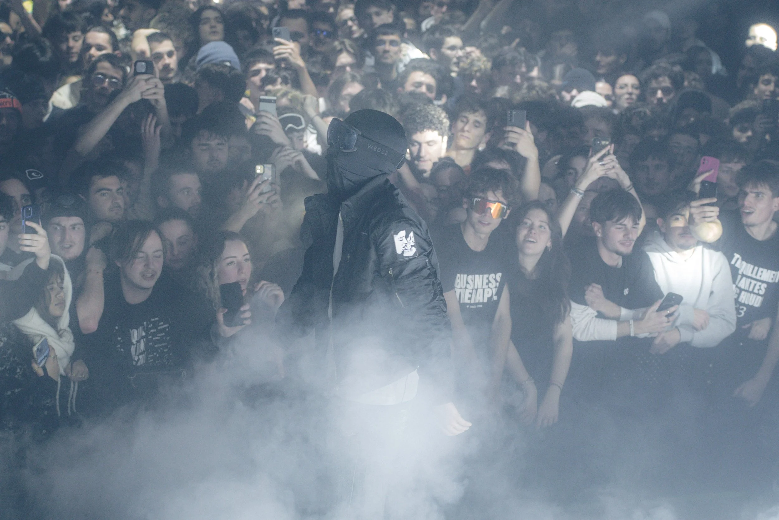 Un homme avec un masque et une veste noire, entouré de nombreuses personnes qui prennent des photos ou vidéos, dans une ambiance sombre avec de la fumée.