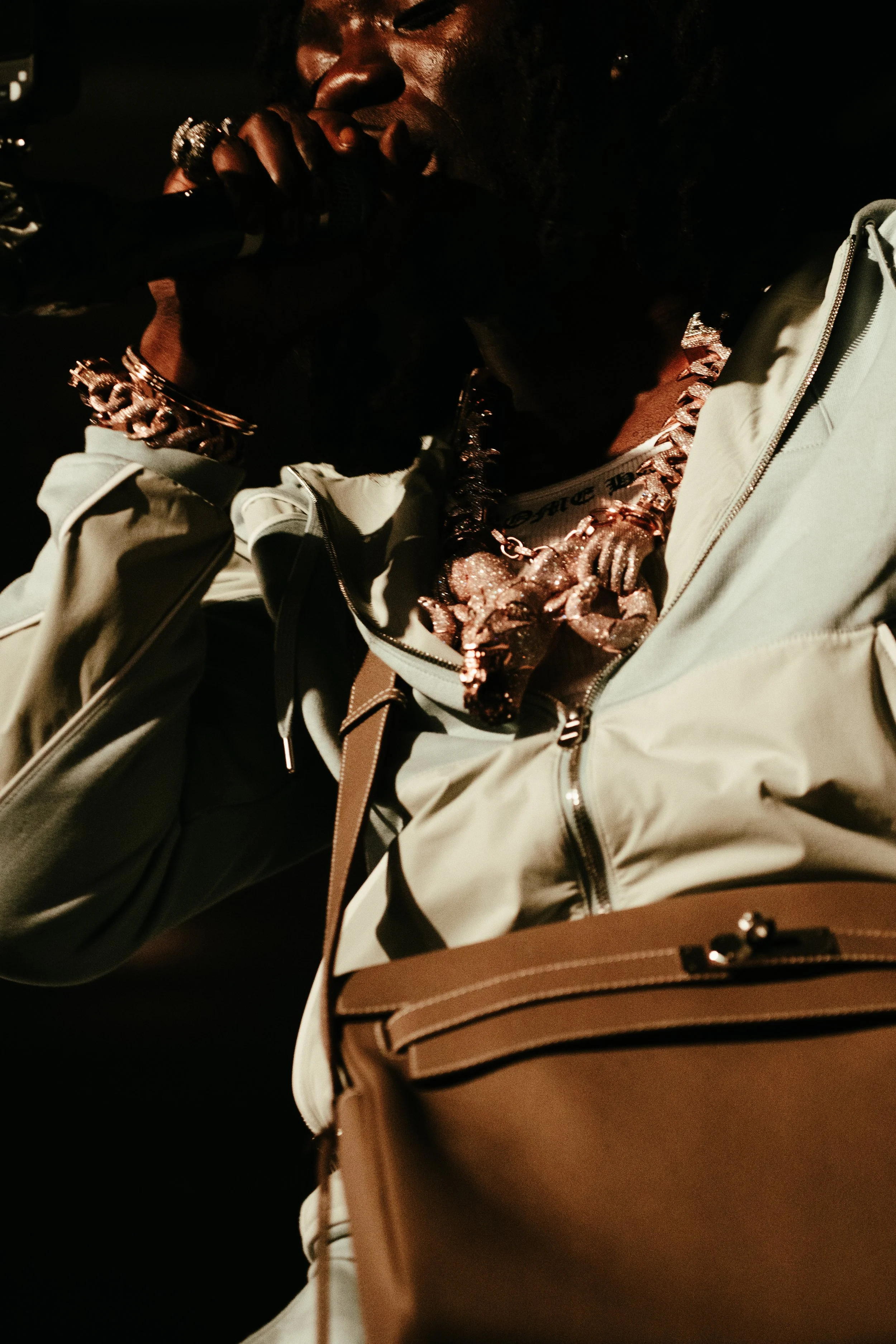 Personne portant plusieurs bijoux en or, tenant un objet noir près de la bouche, avec un sac à dos beige et des vêtements décontractés.