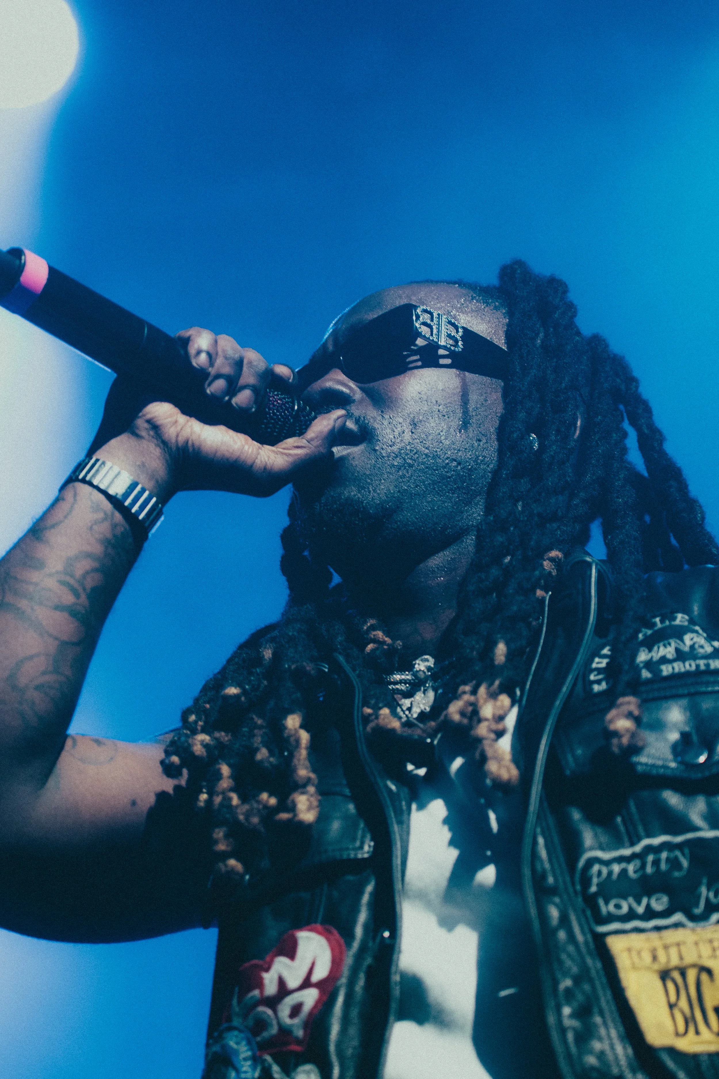 Un homme portant des vêtements en cuir décorés de patches et des lunettes de soleil, chantant ou parlant dans un microphone à haute voix, avec un fond bleu et un éclairage dramatique.