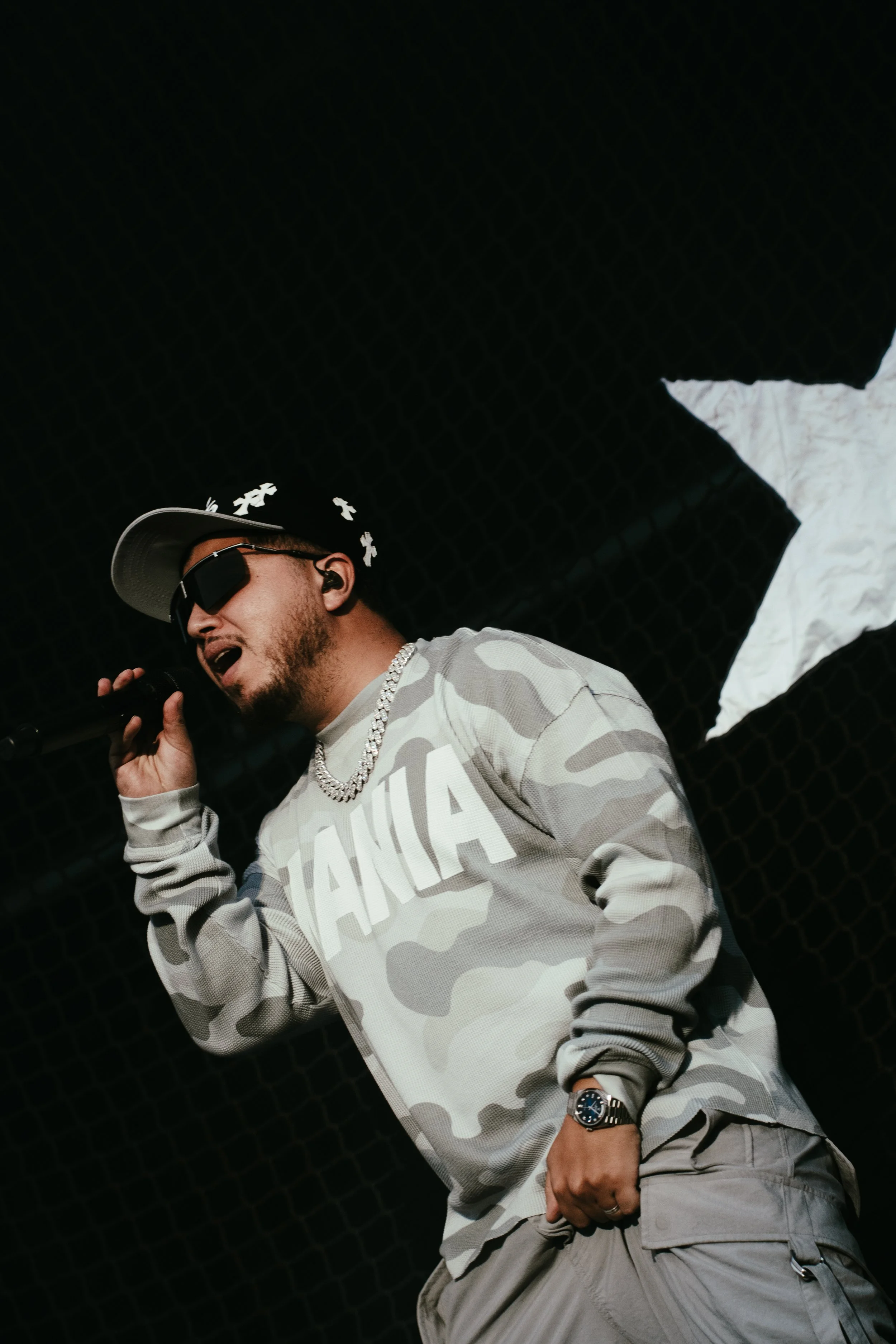 Un homme portant des lunettes de soleil, un chapeau noir et un collier, en train de chanter ou parler en microphone, portant un sweat à motif camouflage avec le mot 'JAHMIA' dessus, dans un décor sombre avec un grand drapeau noir et blanc en arrière-