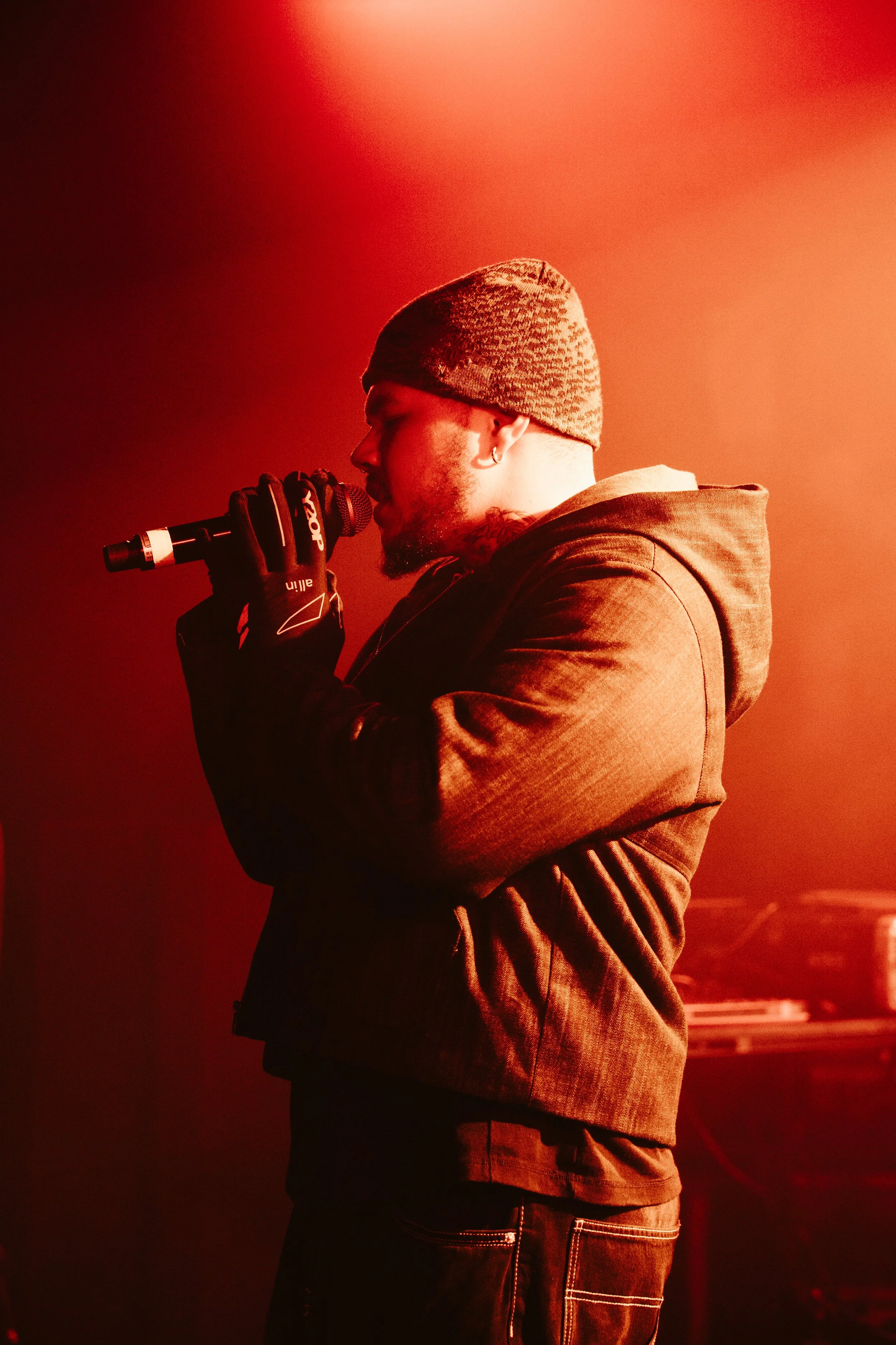 Un homme chante dans un microphone, portant un bonnet et des gants, éclairé par une lumière rouge sur scène.