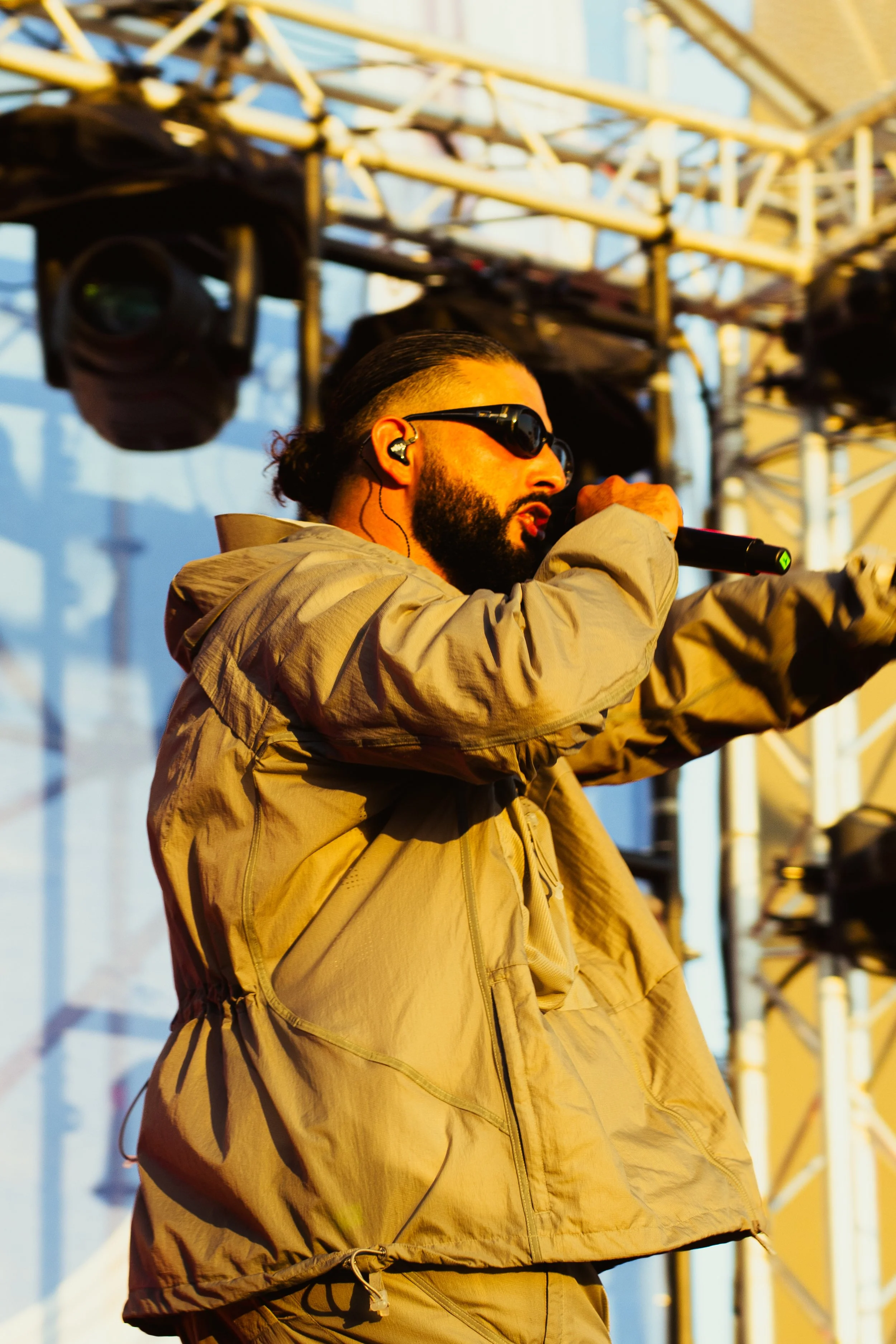 Un homme portant des lunettes de soleil et un casque d'écoute chante dans un microphone lors d'un concert en plein air, avec une structure de scène en métal en arrière-plan.