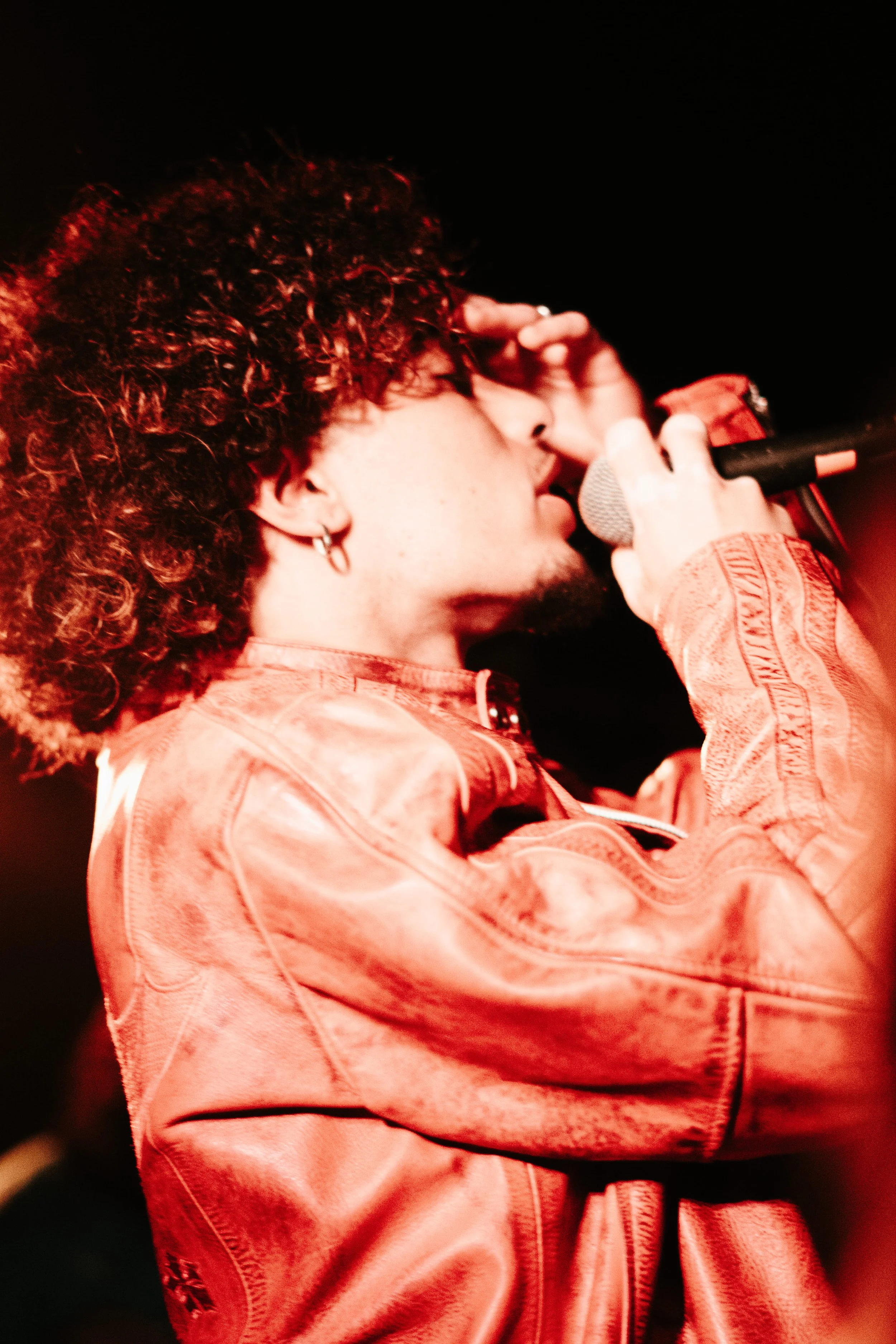 Un homme avec des cheveux bouclés portant une veste en cuir tient un microphone lors d'une performance ou d'un concert, dans un environnement sombre et éclairé par une lumière rouge.