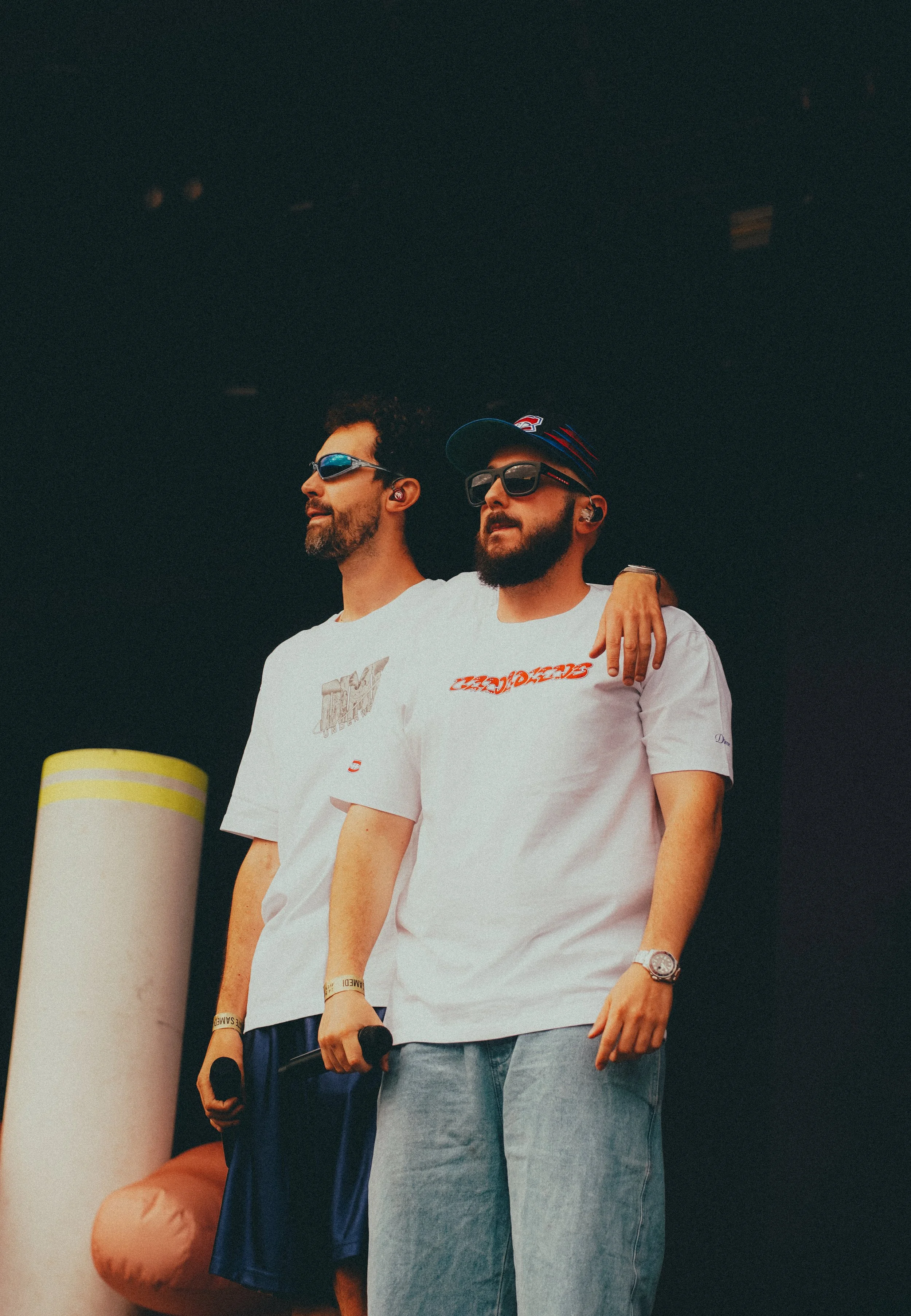 Deux jeunes hommes portant des T-shirts blancs et des lunettes de soleil, l'un d'eux avec une casquette, debout côte à côte sur scène, avec une personne à l'arrière-plan à gauche.