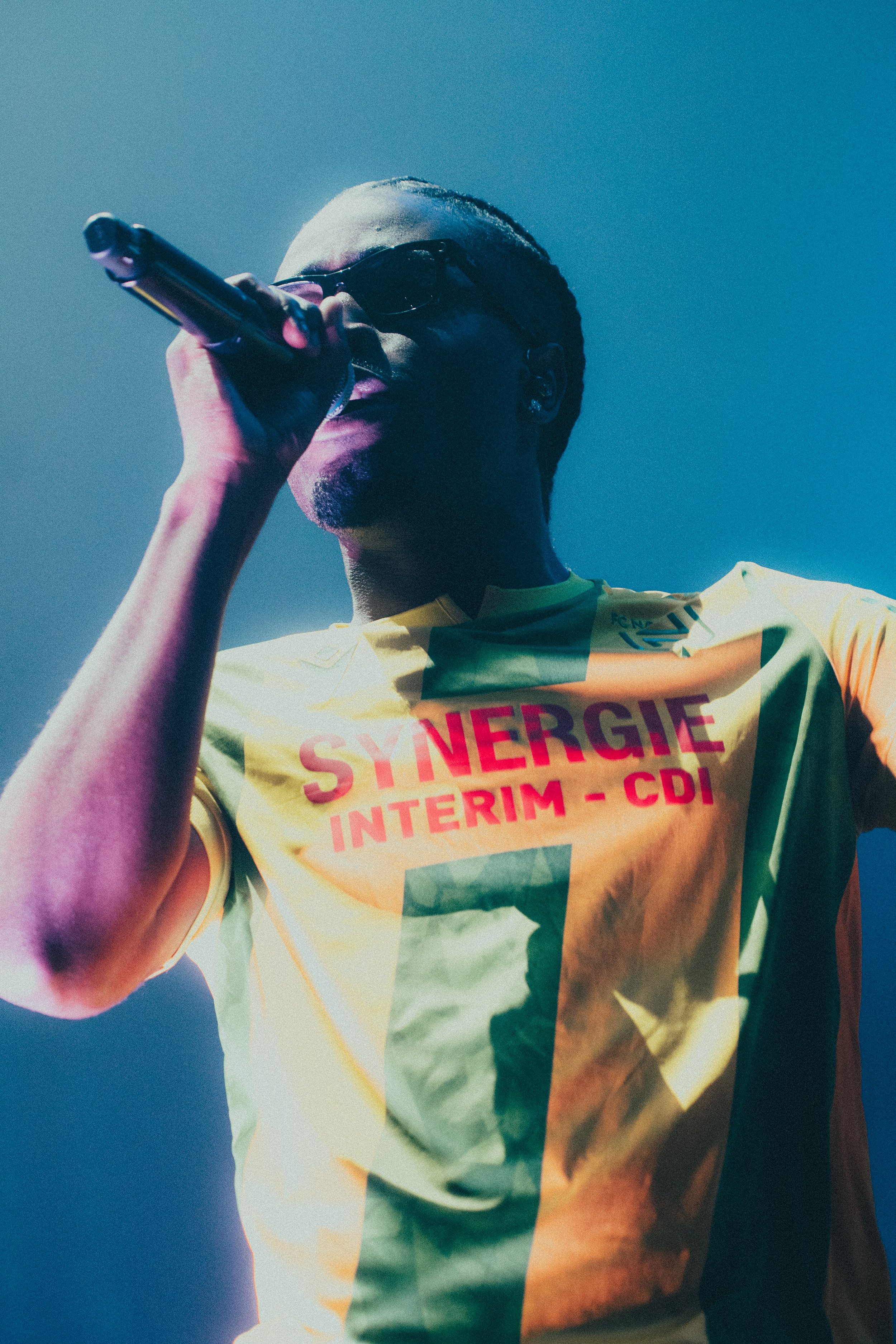 Un chanteur portant des lunettes noires, vêtu d'un maillot jaune avec l'inscription "SYNERGIE INTERIM - CDI" en rouge, chante dans un microphone sur scène avec un éclairage bleu.