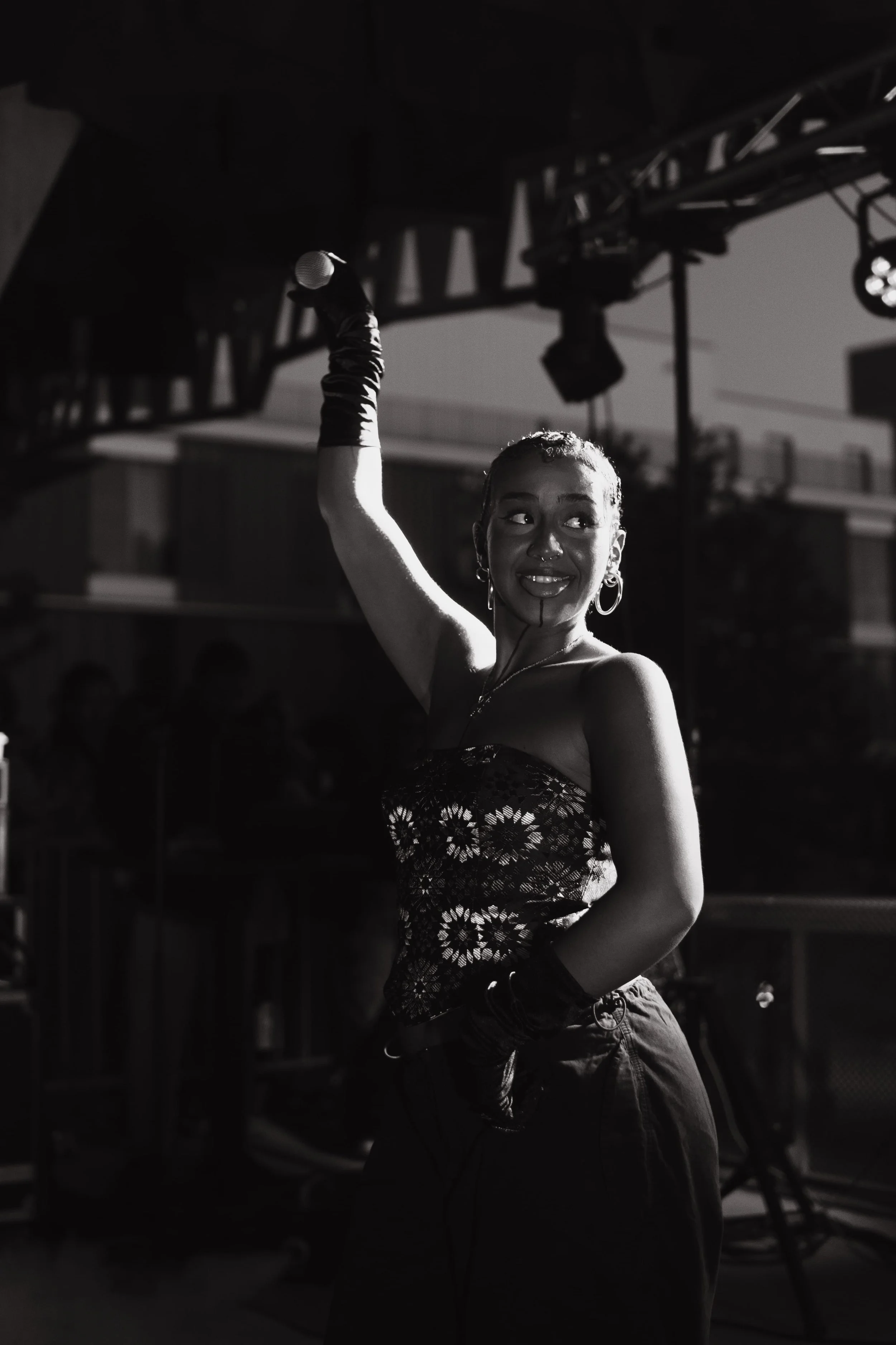 Une femme souriante, portant une robe strapless et des accessoires, tenant un microphone en l'air lors d'une performance sur scène, en noir et blanc.