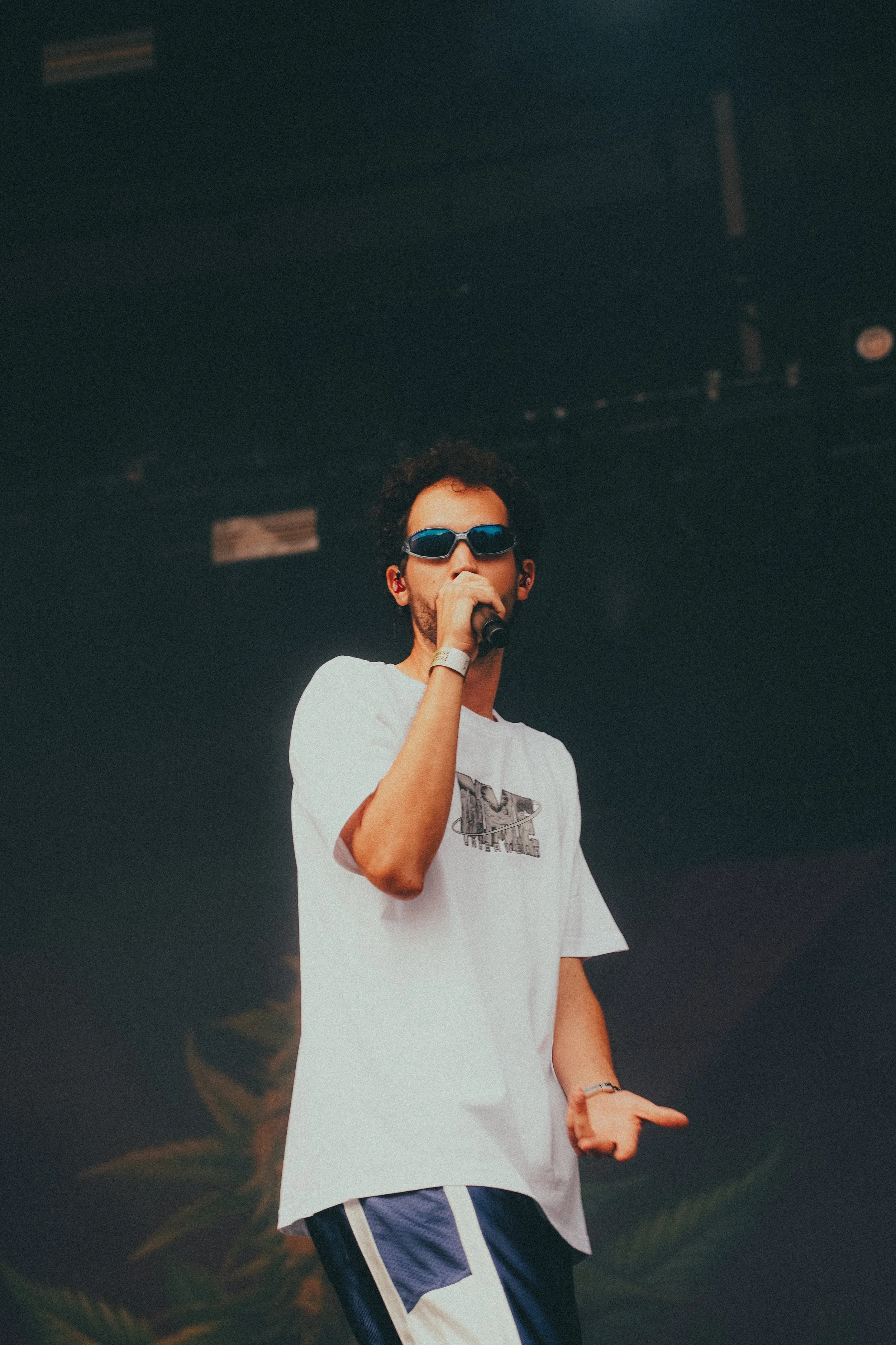 Un jeune homme portant des lunettes de soleil noires et un t-shirt blanc avec un logo, tenant un microphone, sur scène lors d'une performance.