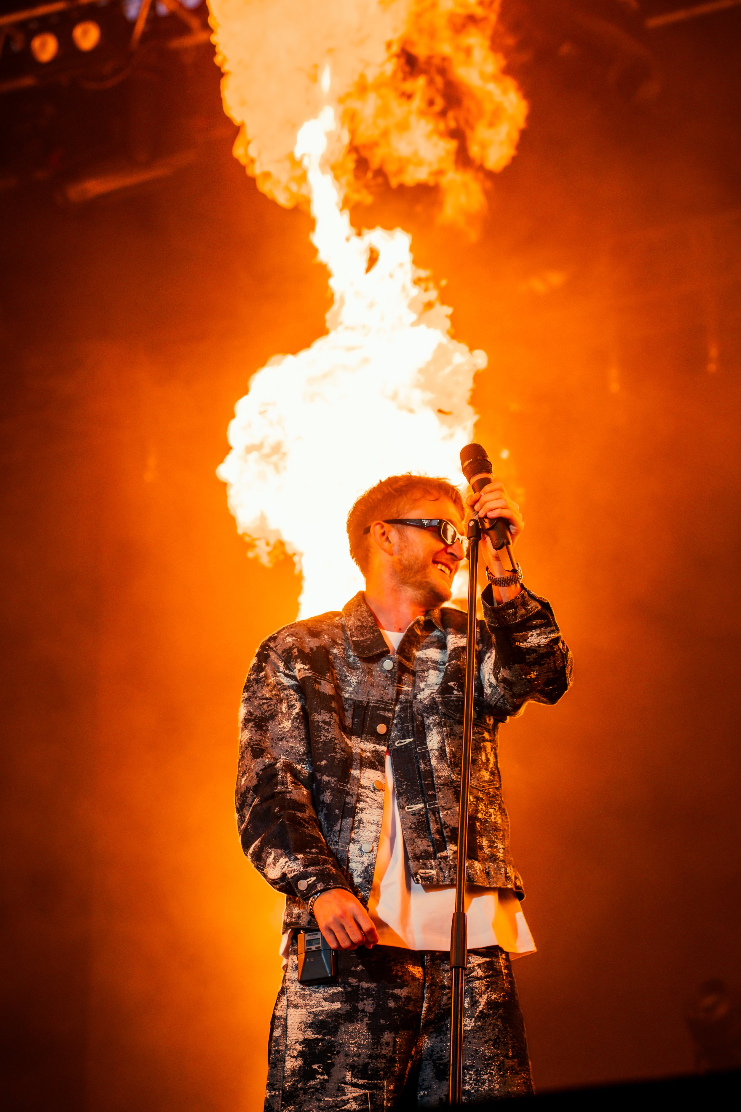 Un homme portant des vêtements de style camouflage, portant des lunettes de soleil, tenant un microphone et se trouvant devant un feu d'artifice en forme de flammes lors d'un spectacle sur scène.