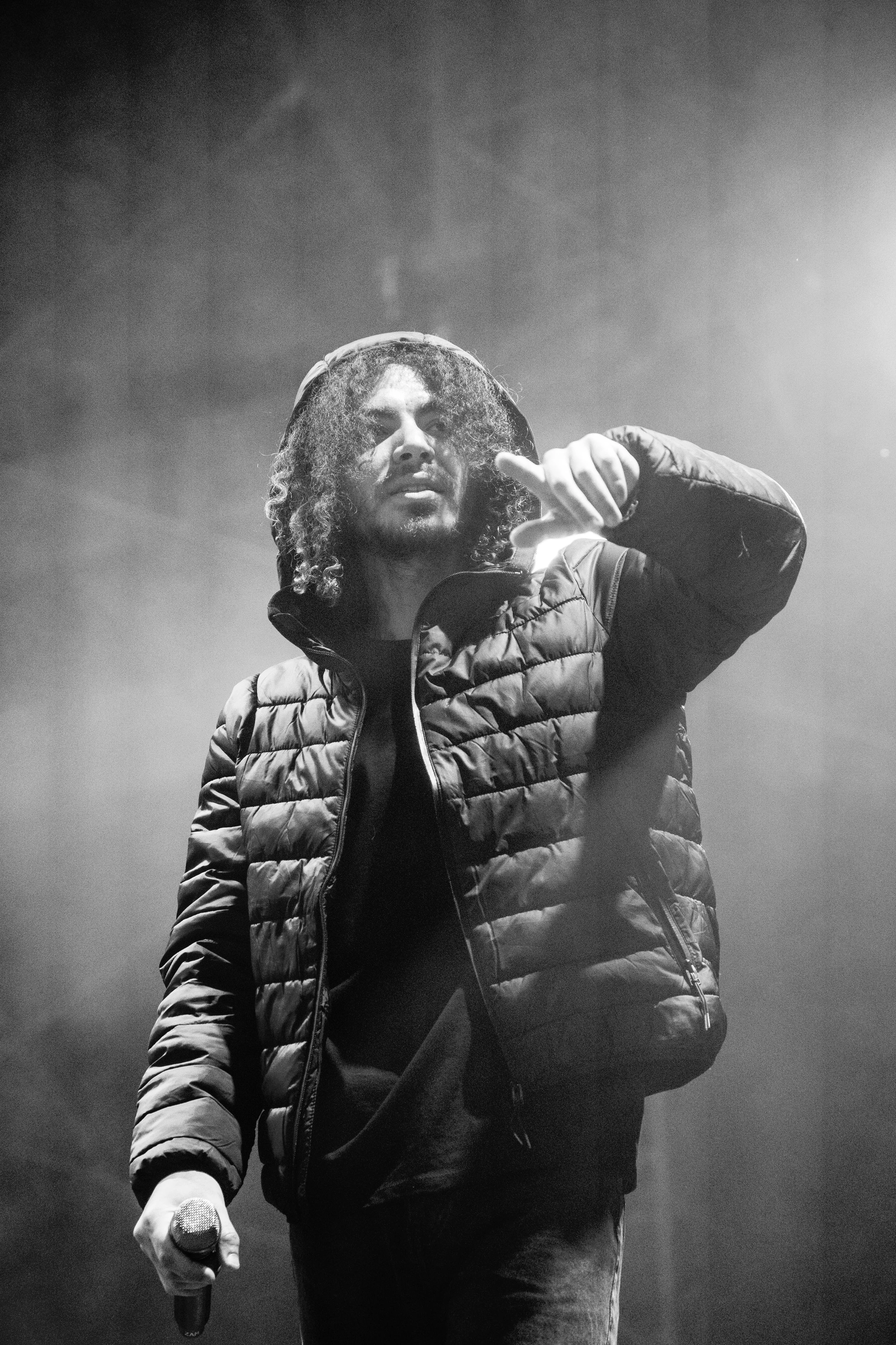 Un homme avec des cheveux bouclés longs, portant une veste rembourrée, tenant un microphone, sur scène avec des lumières et de la fumée.