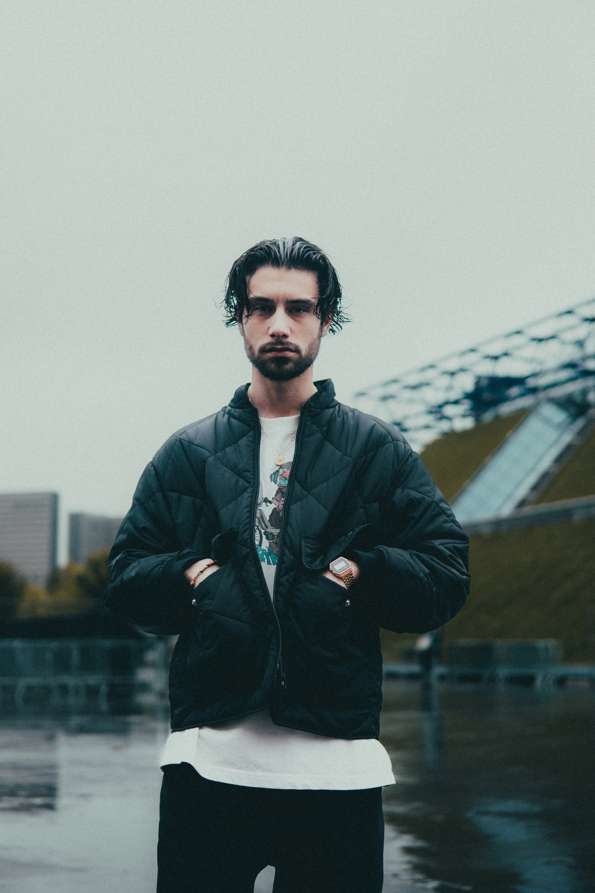 Un homme avec des cheveux noirs et une barbe porte une veste noire et regarde vers l'objectif, dans un environnement urbain par temps nuageux.