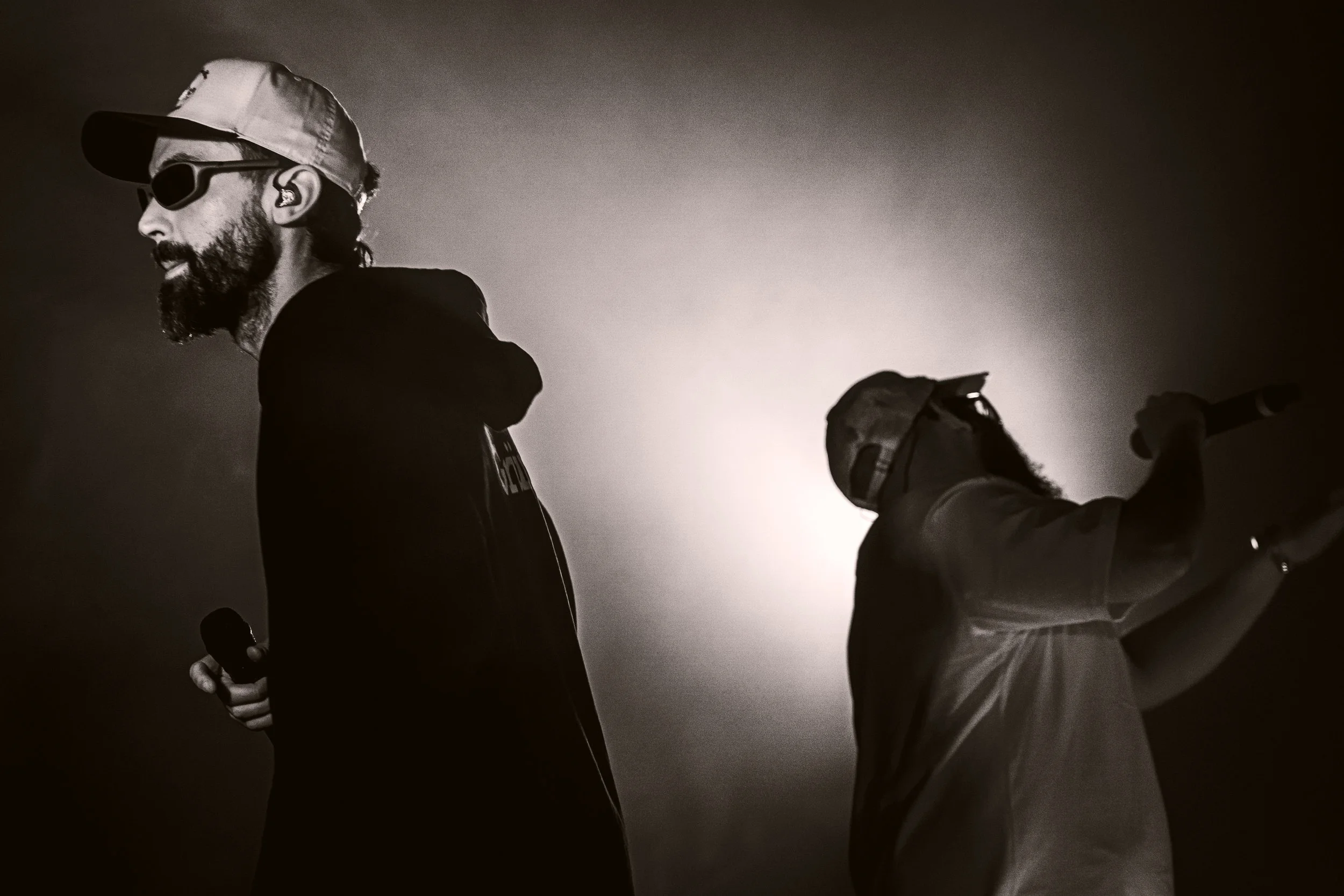 Deux hommes debout, un avec un micro dans la main, en noir et blanc, avec un éclairage de derrière.