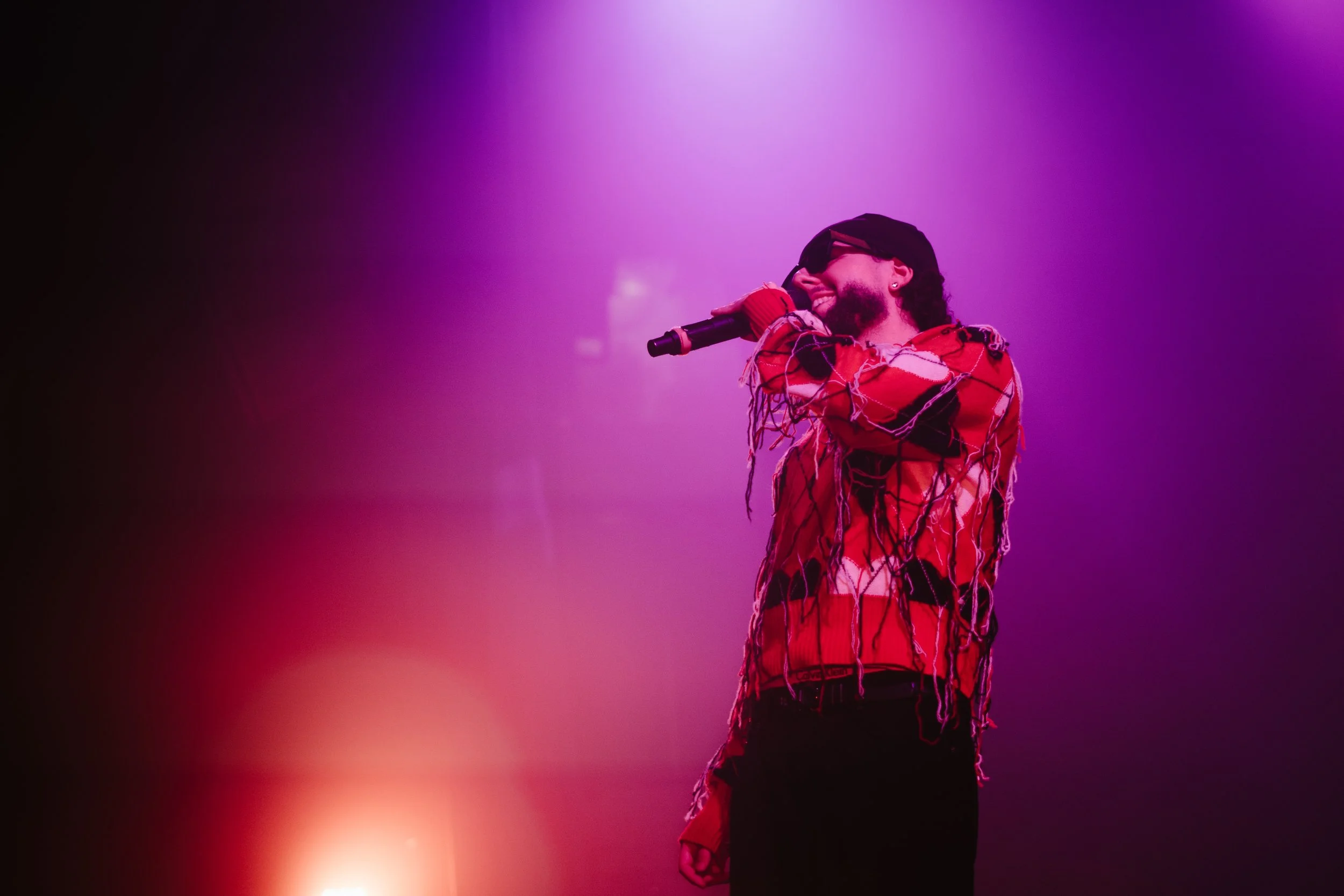 Chanteur avec un micro sur scène, vêtu d’un vêtement rouge déchiré, sous des lumières violettes et roses.