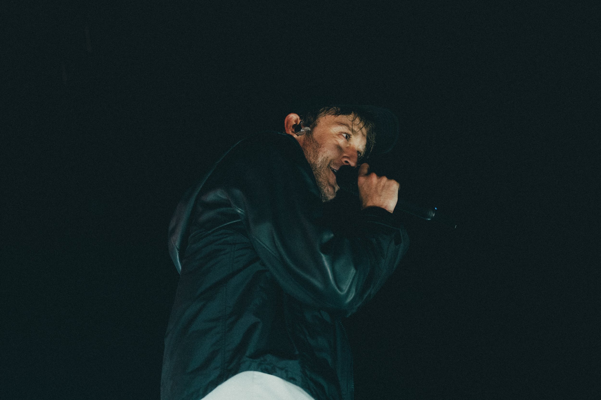 Un homme portant une veste en cuir noire, un casque, et tenant un microphone, chantant ou parlant dans l'obscurité.