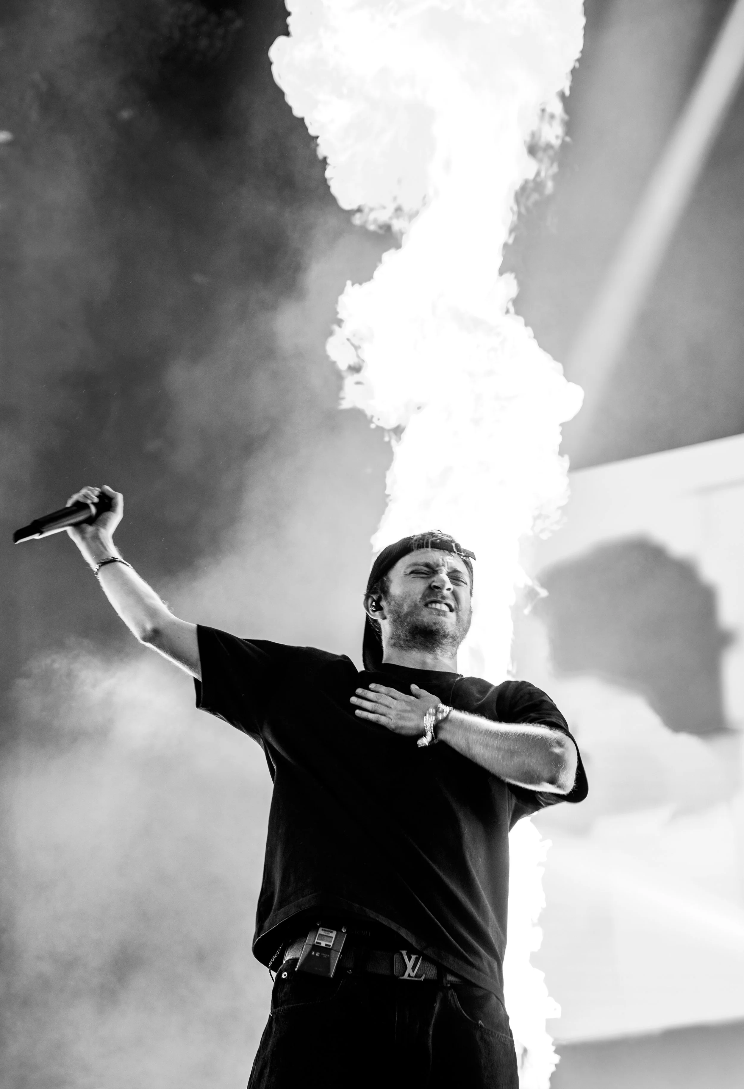 Un homme lors d'un concert, il tient un micro dans sa main droite, la main gauche sur la poitrine, avec une expression intense, et un effet de feu ou de flammes derrière lui.