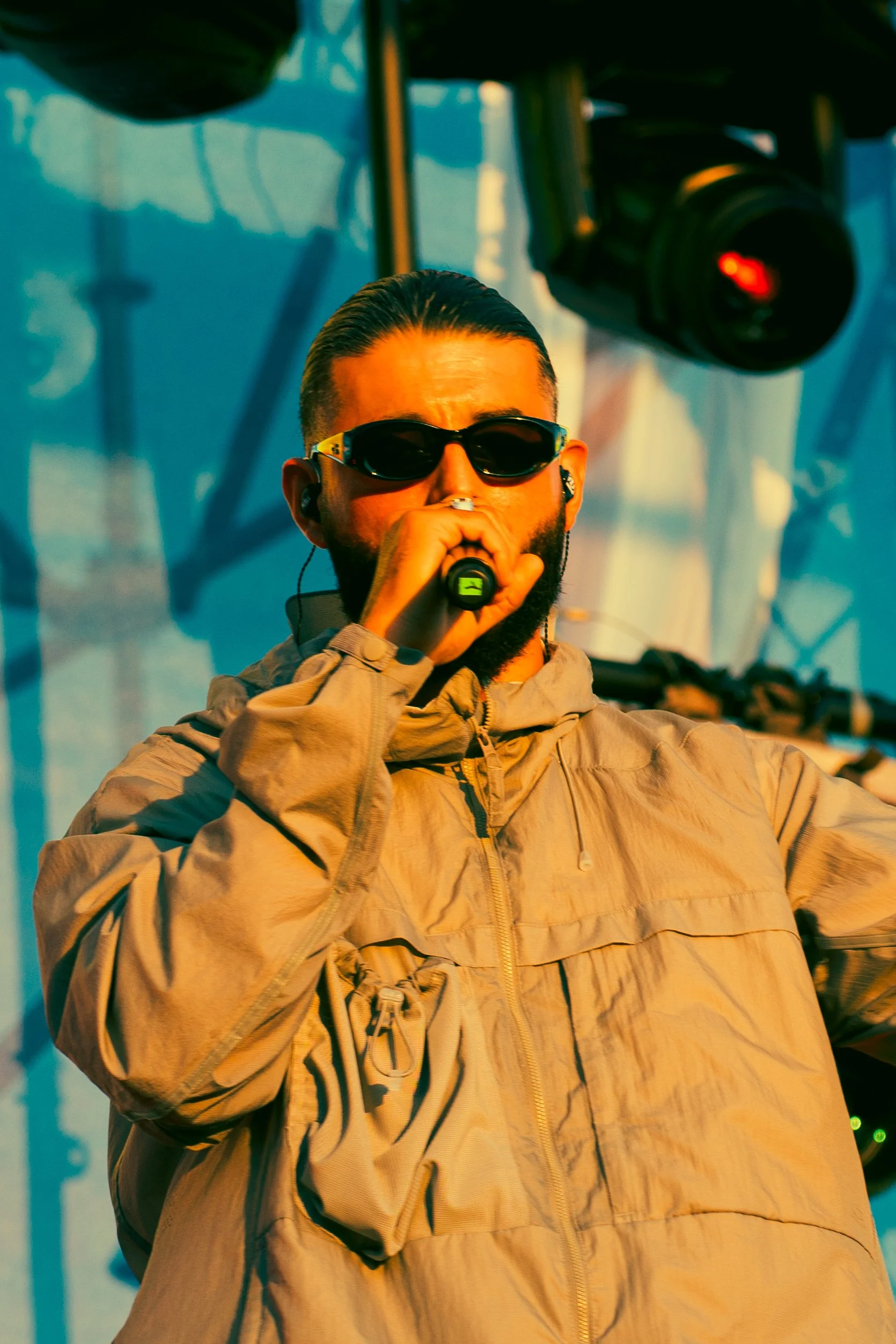 Un homme portant des lunettes de soleil noires et un casque d'écoute, en train de chanter ou parler dans un microphone lors d'un spectacle ou événement en extérieur.