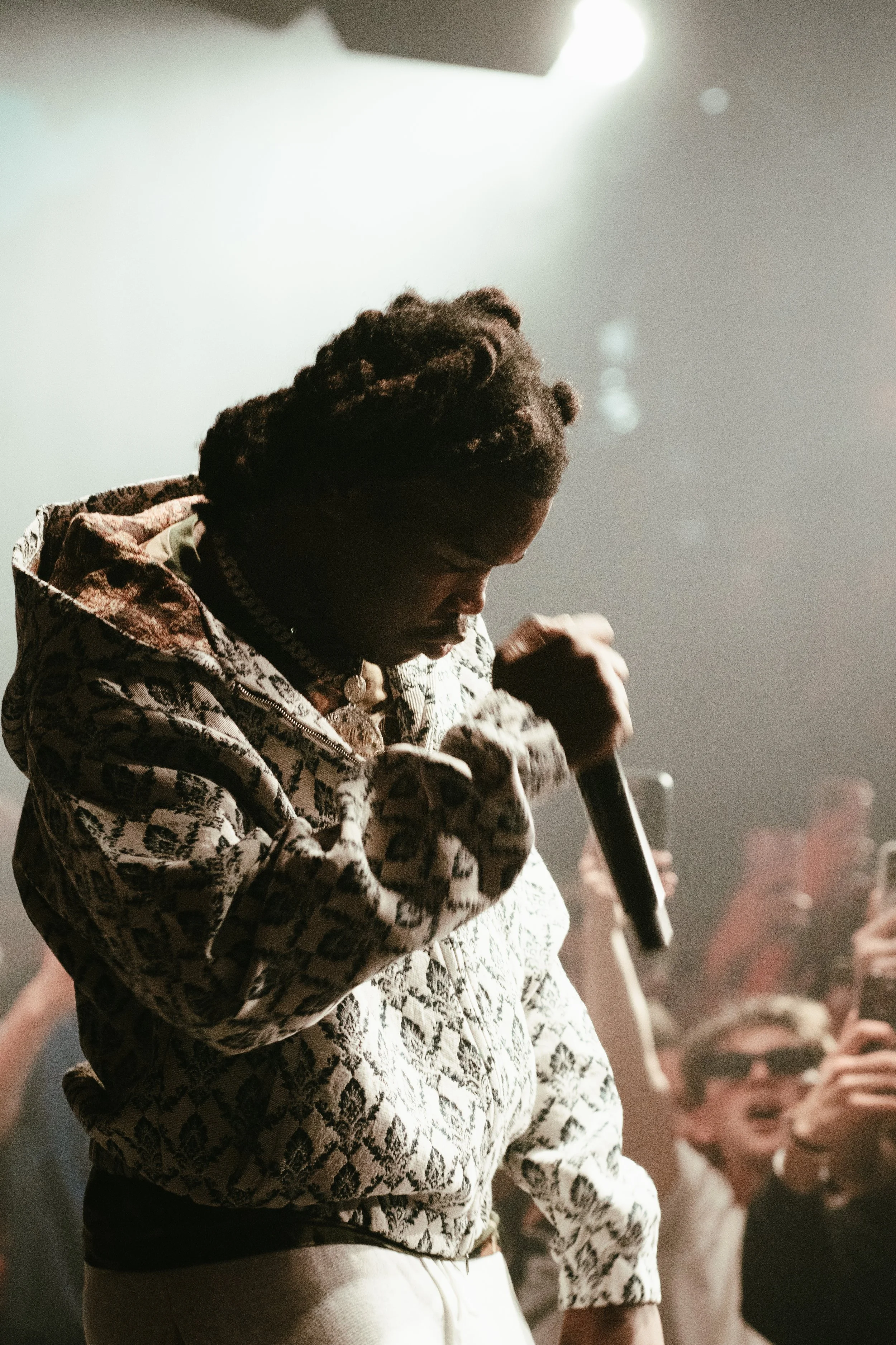 Homme avec dreadlocks portant un hoodie à motif, tenant un microphone lors d'une performance devant un public.
