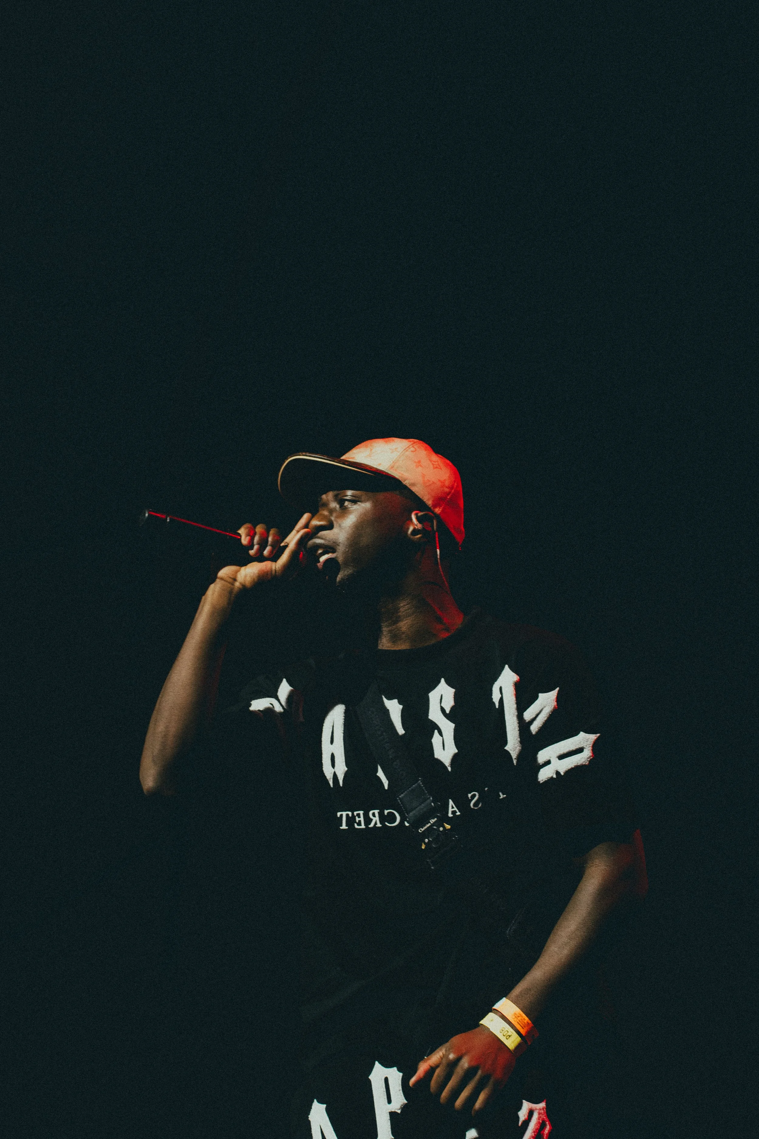 Un homme tenant un microphone, portant un chapeau de baseball rouge et noir, en train de chanter ou parler, dans un environnement sombre.