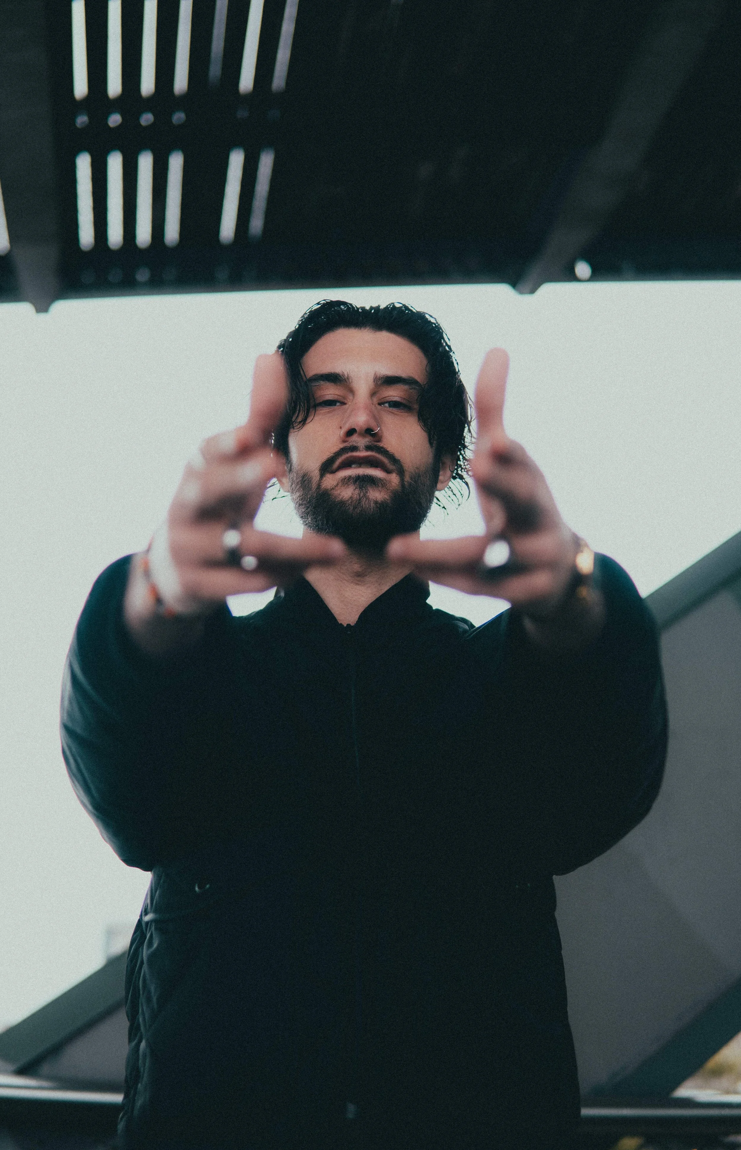 Un homme avec des cheveux noirs, barbe et un piercing, regardant vers le bas en pointant ses doigts vers la caméra, avec un ciel gris en arrière-plan.