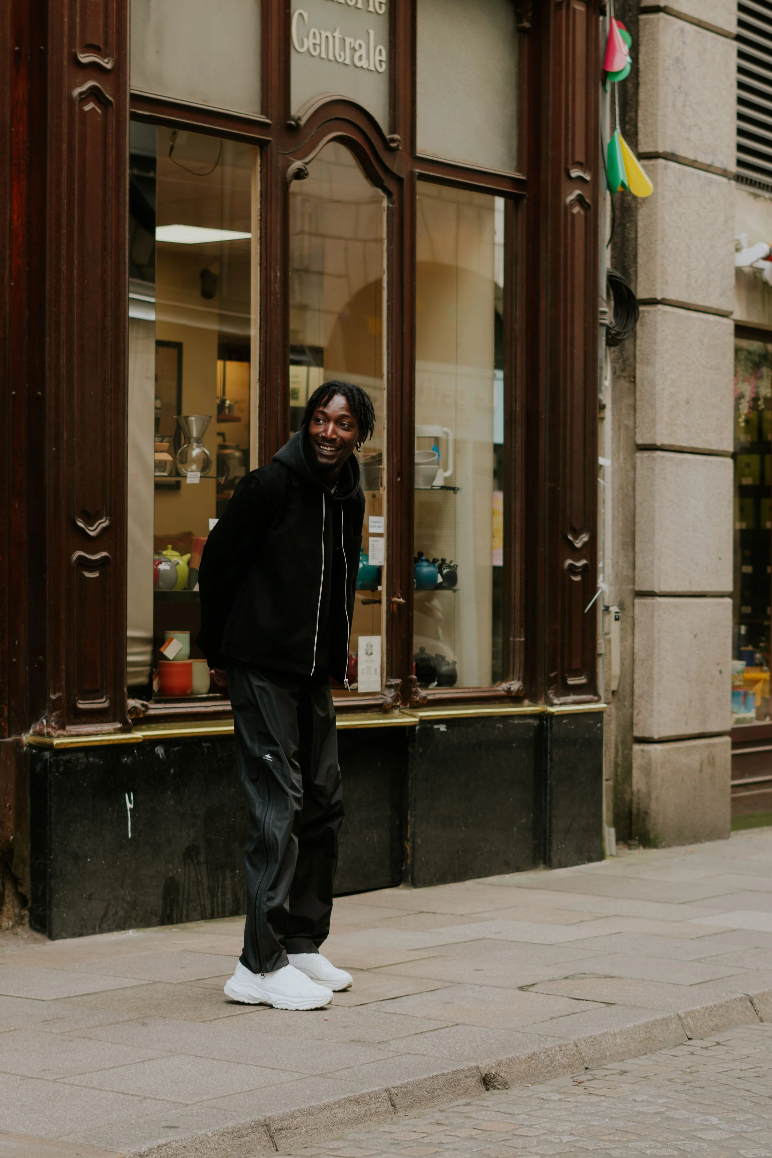 Un homme souriant debout sur un trottoir devant une vitrine de magasin avec des objets en céramique, portant des vêtements de sport noirs et des baskets blanches.
