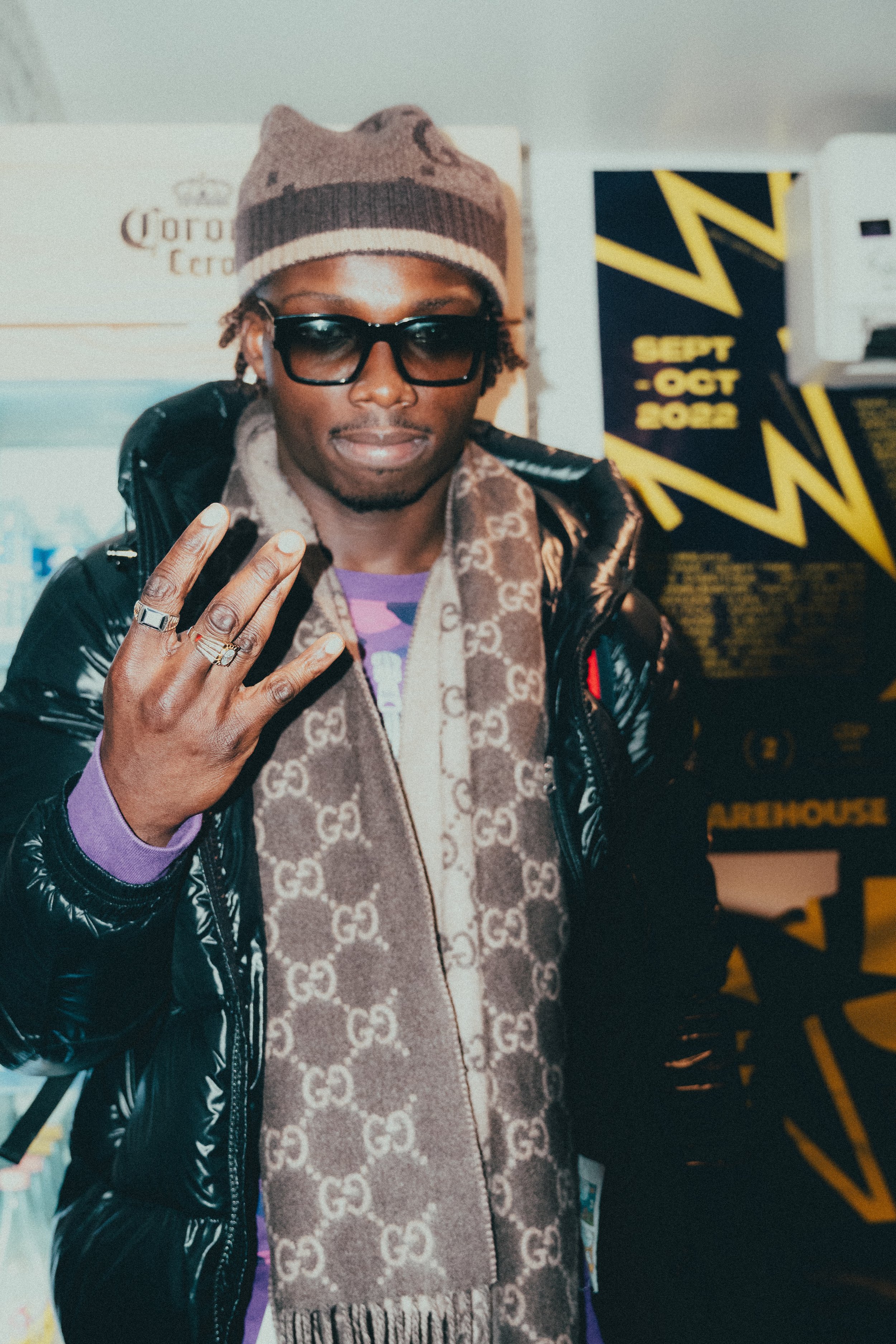 Homme portant une écharpe Gucci, lunettes de soleil noires, chapeau beige, veste noire en PVC, indiquant le chiffre trois avec sa main, en arrière-plan un panneau avec des dates et un éclair.