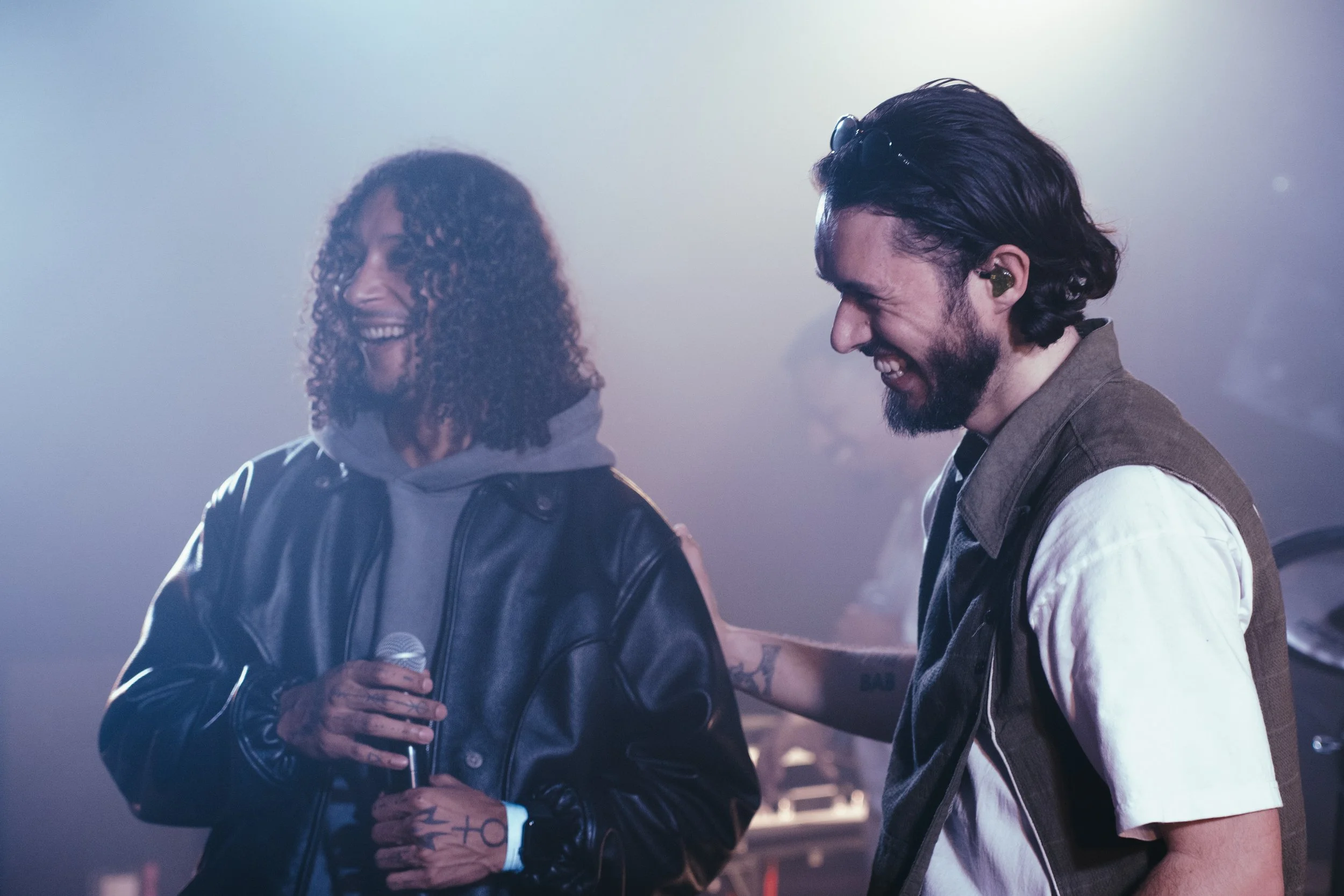 Deux personnes souriantes se regardent lors d'une performance ou d'une fête, l'une d'elles tient un micro, ambiance lumineuse tamisée.