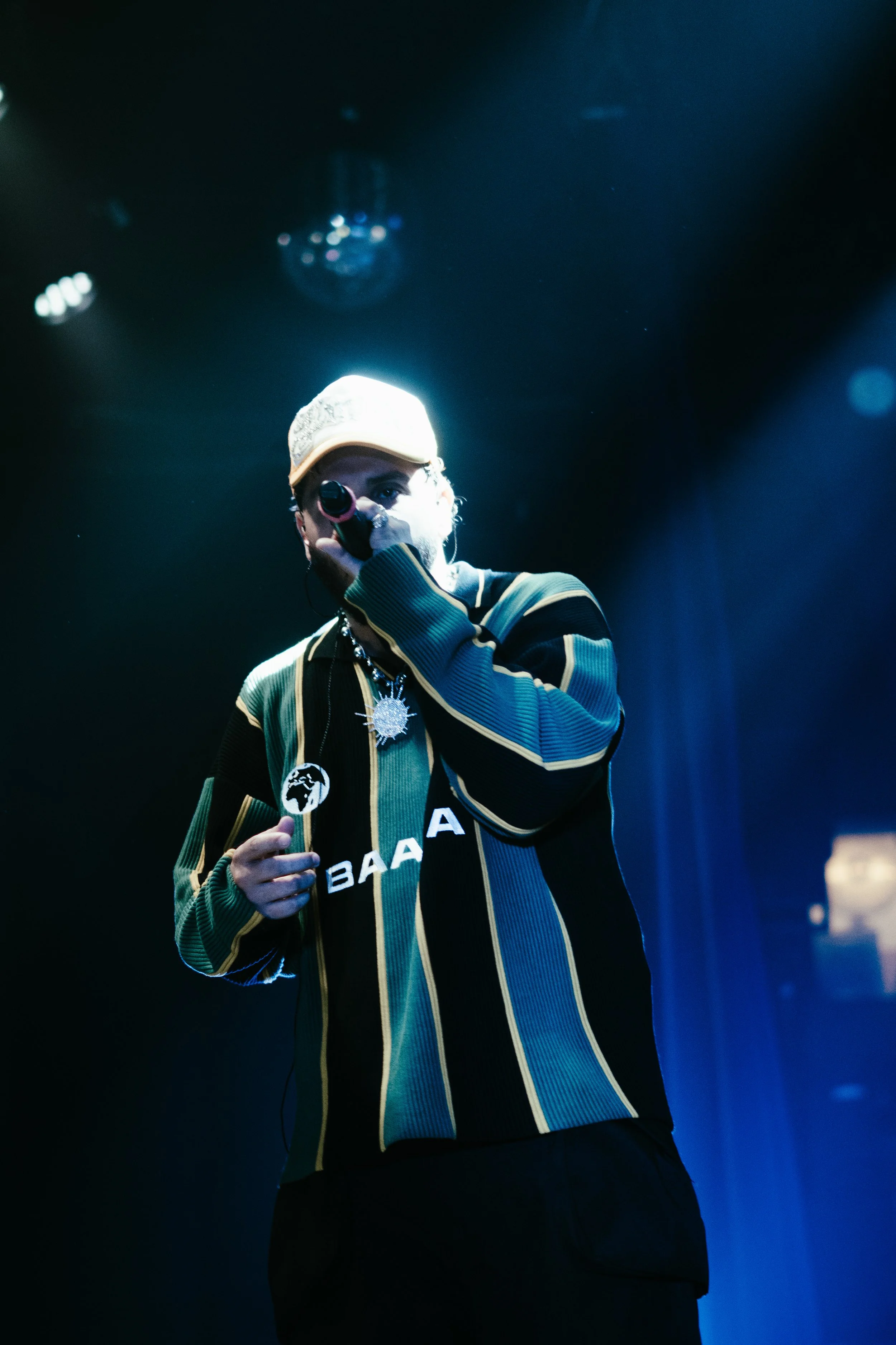 Un homme en tenue de scène satisfait sur scène, micros et lumière bleue, portant un bonnet beige, un sweat à rayures vertes, noires et dorées, avec des bijoux, lors d'une performance.