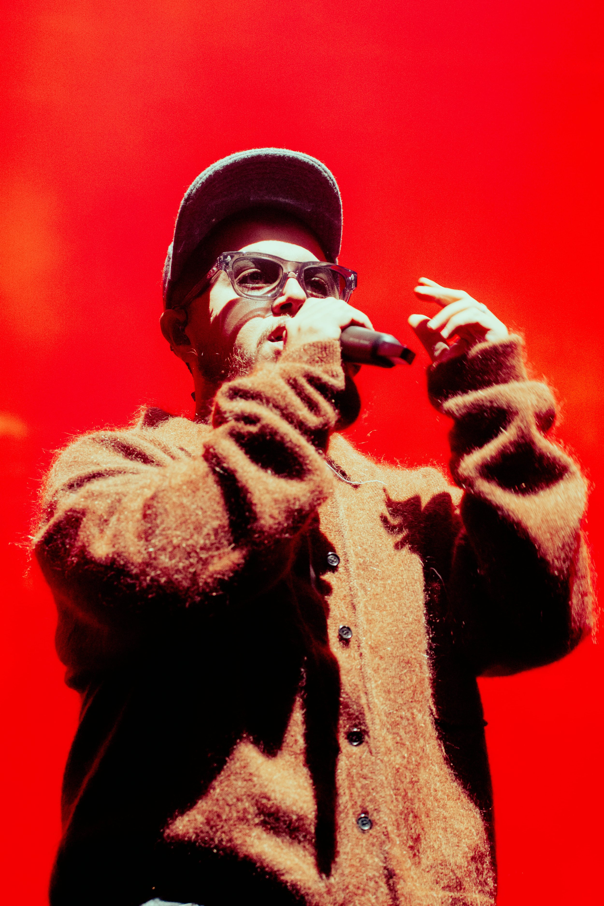 Un homme portant des lunettes de soleil, une casquette, un sweat à motif camouflage, et une chaîne en/avec un collier en or, chantant dans un micro.