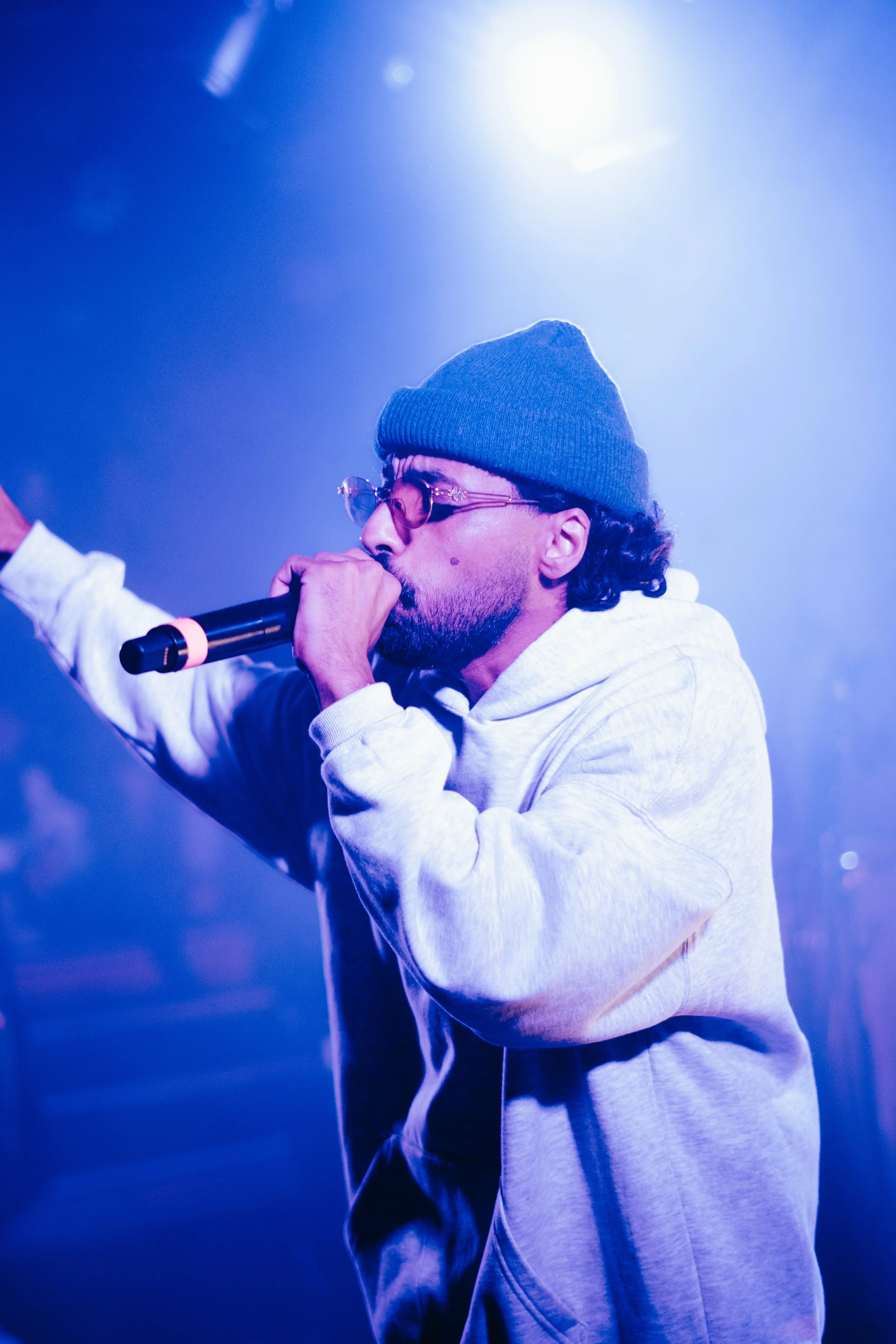 Homme portant un bonnet gris, lunettes, et sweat blanc, chantant avec un micro dans une scène de concert avec lumières bleues.