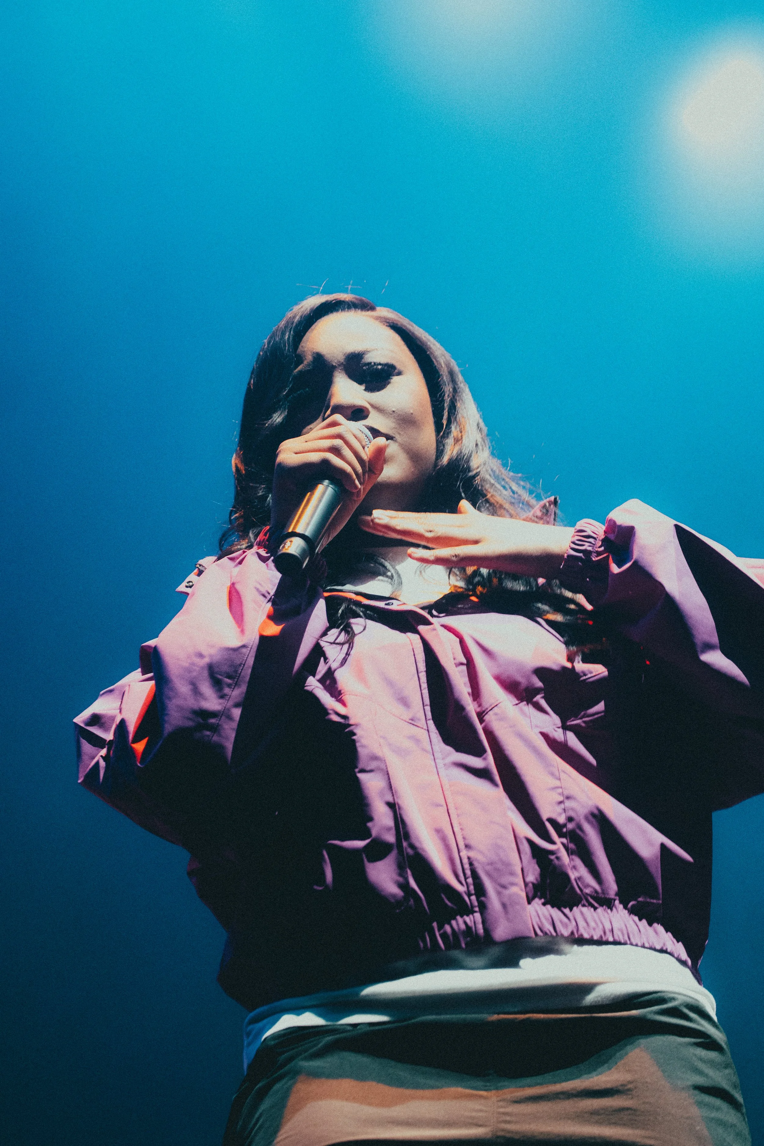 Une femme chante dans un micro, vêtue d'une veste violette, avec un fond de ciel bleu.