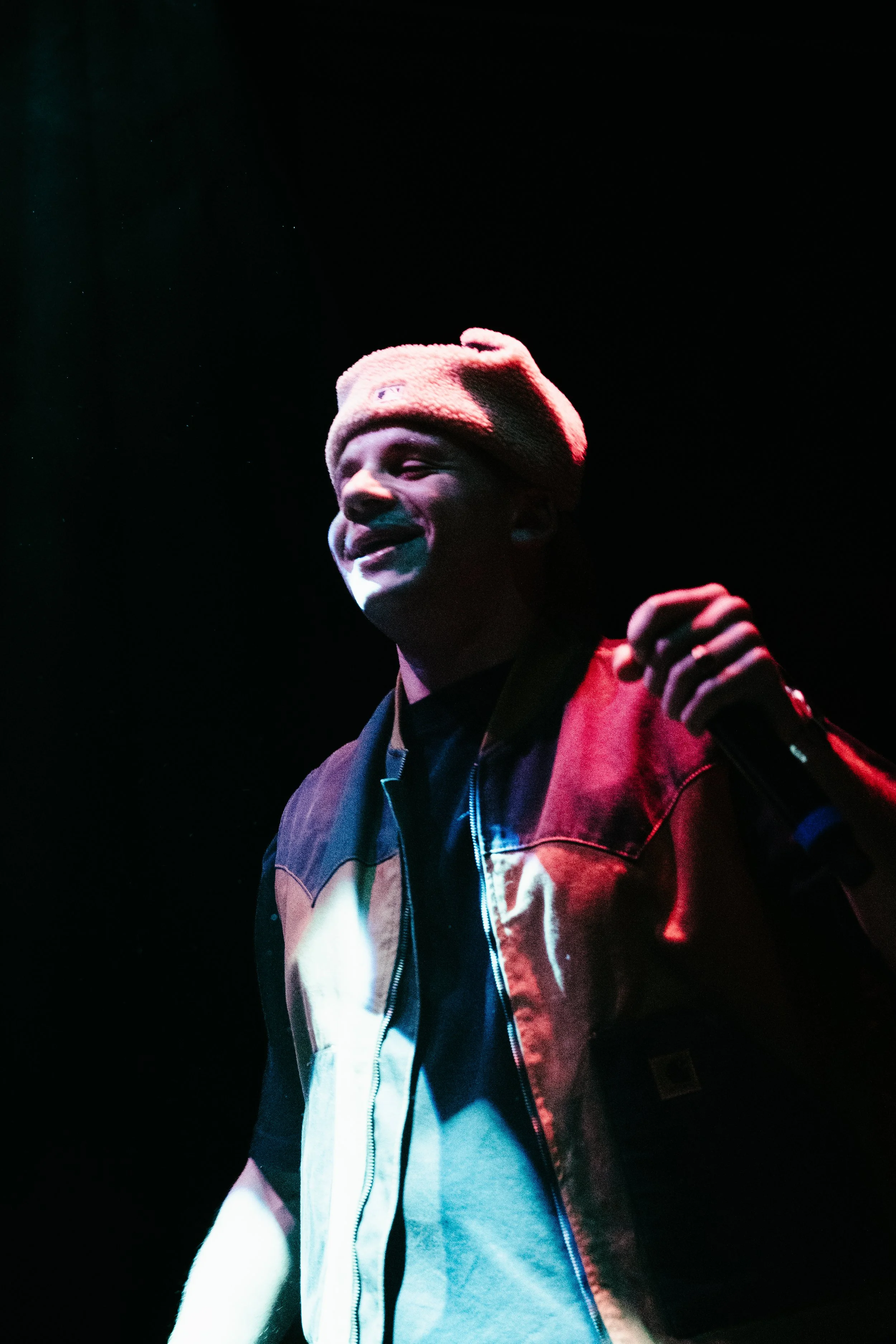 Un jeune homme souriant, portant un bonnet beige et un blouson noir, tenant un microphone dans la main, lors d'une performance sur scène avec un éclairage coloré