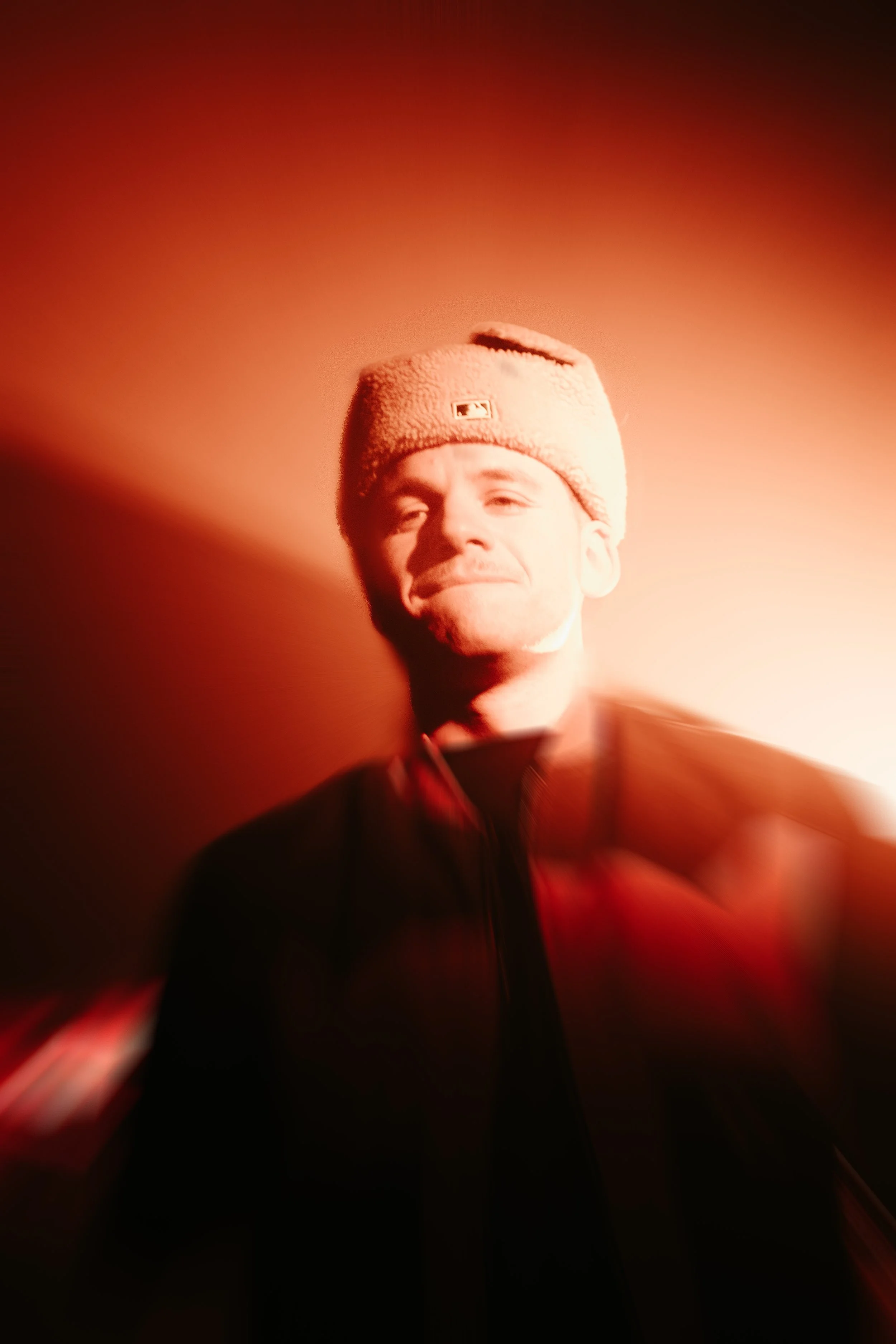 Un homme portant un bonnet beige, souriant légèrement, avec un arrière-plan rougeâtre et un effet de flou.