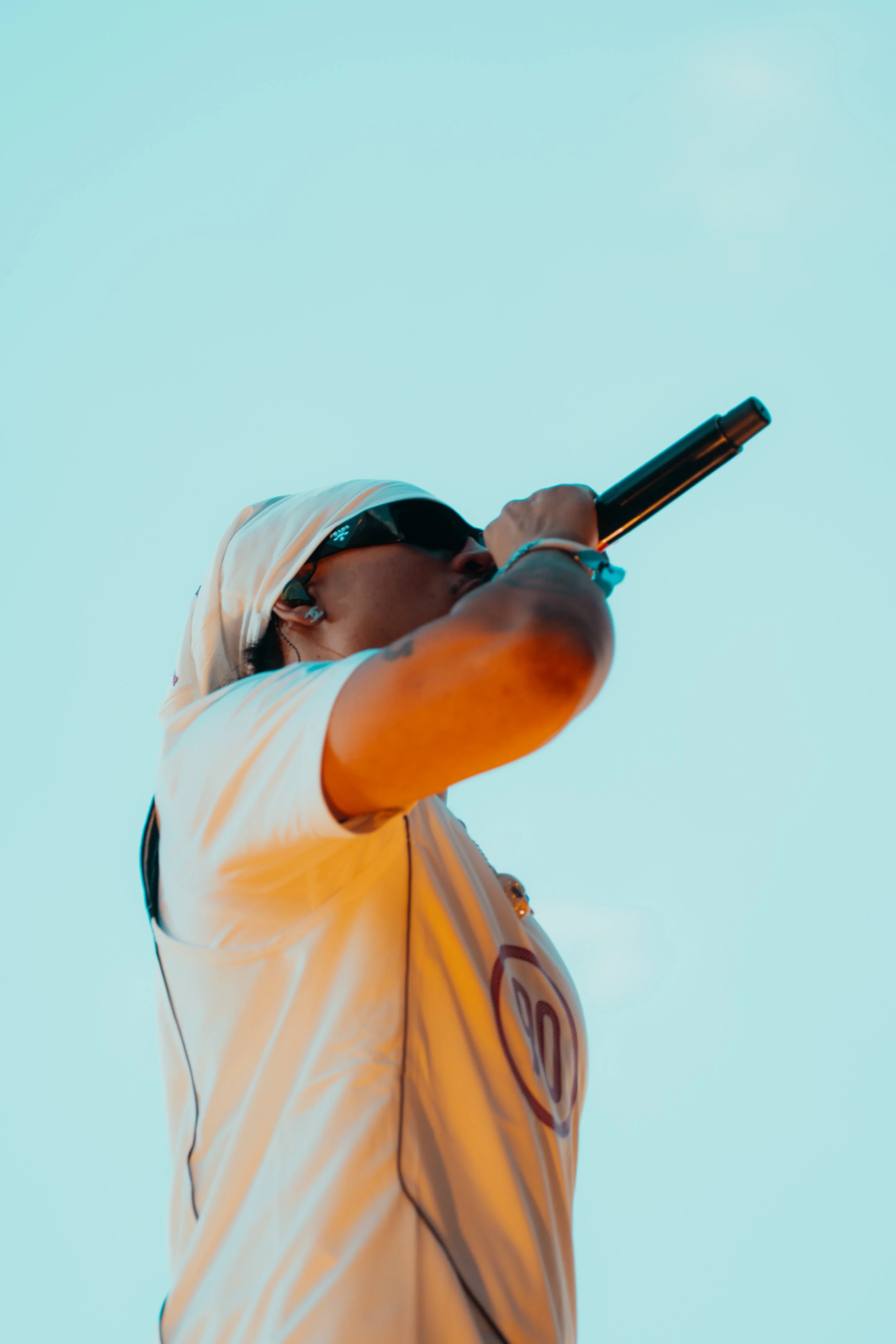 Un homme portant un t-shirt blanc, un bandana blanc sur la tête et des lunettes de soleil chante dans un microphone, vu en contre-plongée contre un ciel clair.