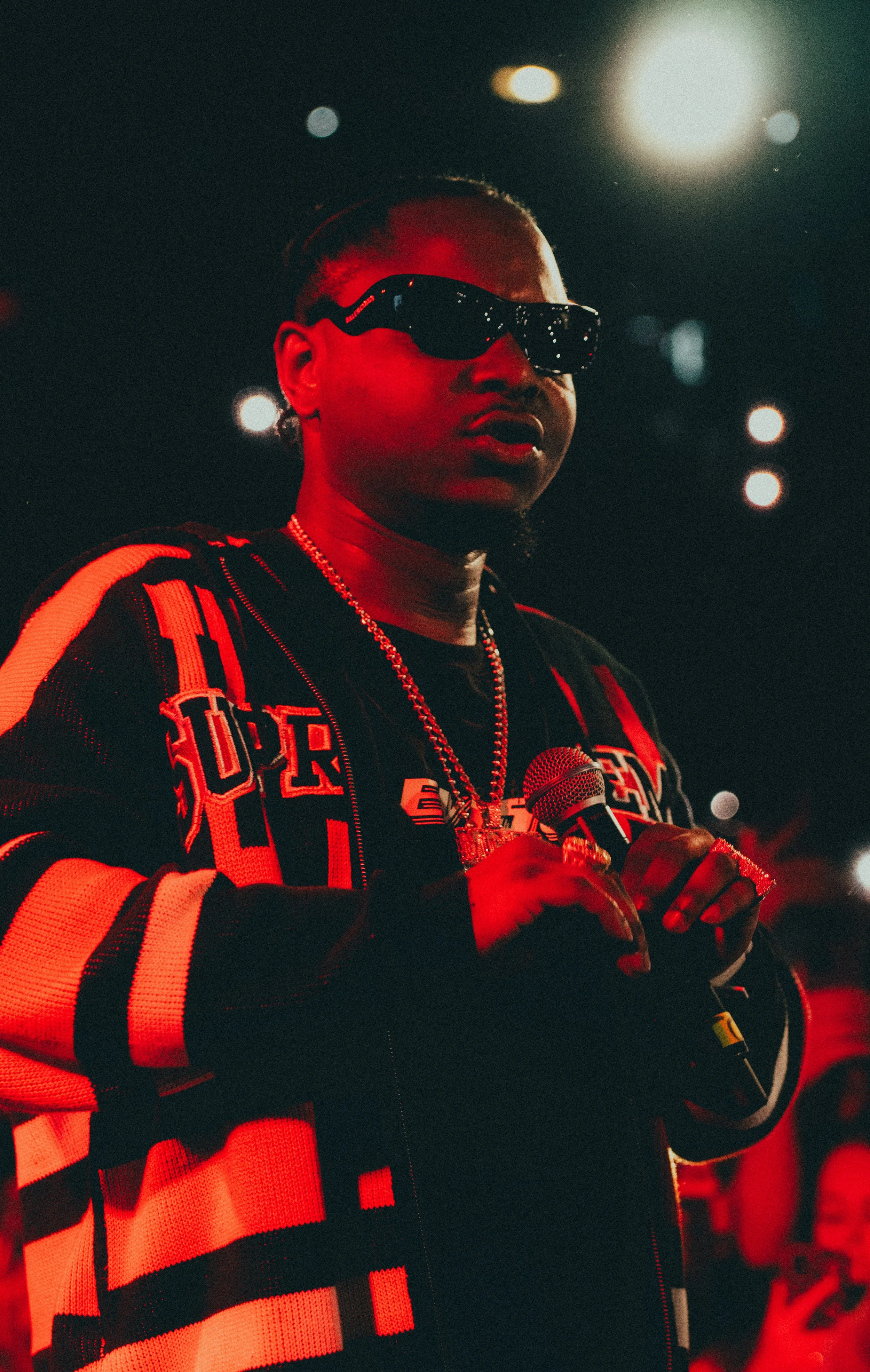 Personne portant des lunettes de soleil noires, un chandail à rayures rouges et noires, tenant un microphone, dans un environnement sombre avec des lumières.