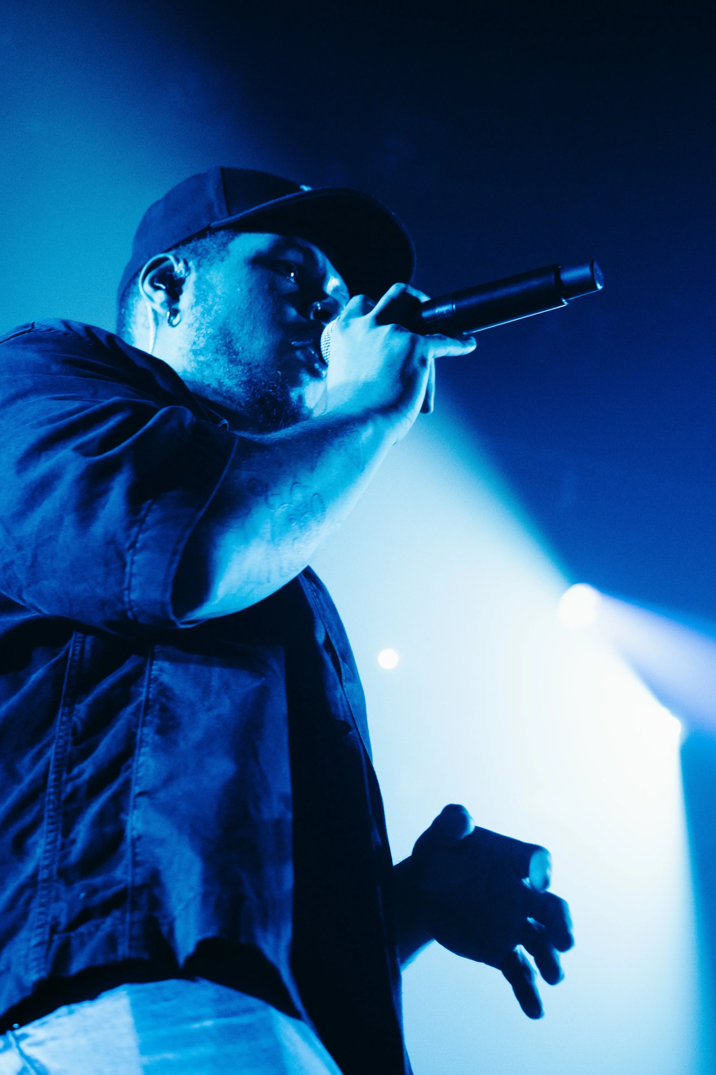 Un homme chante ou parle dans un micro lors d'une performance, avec un éclairage bleu.