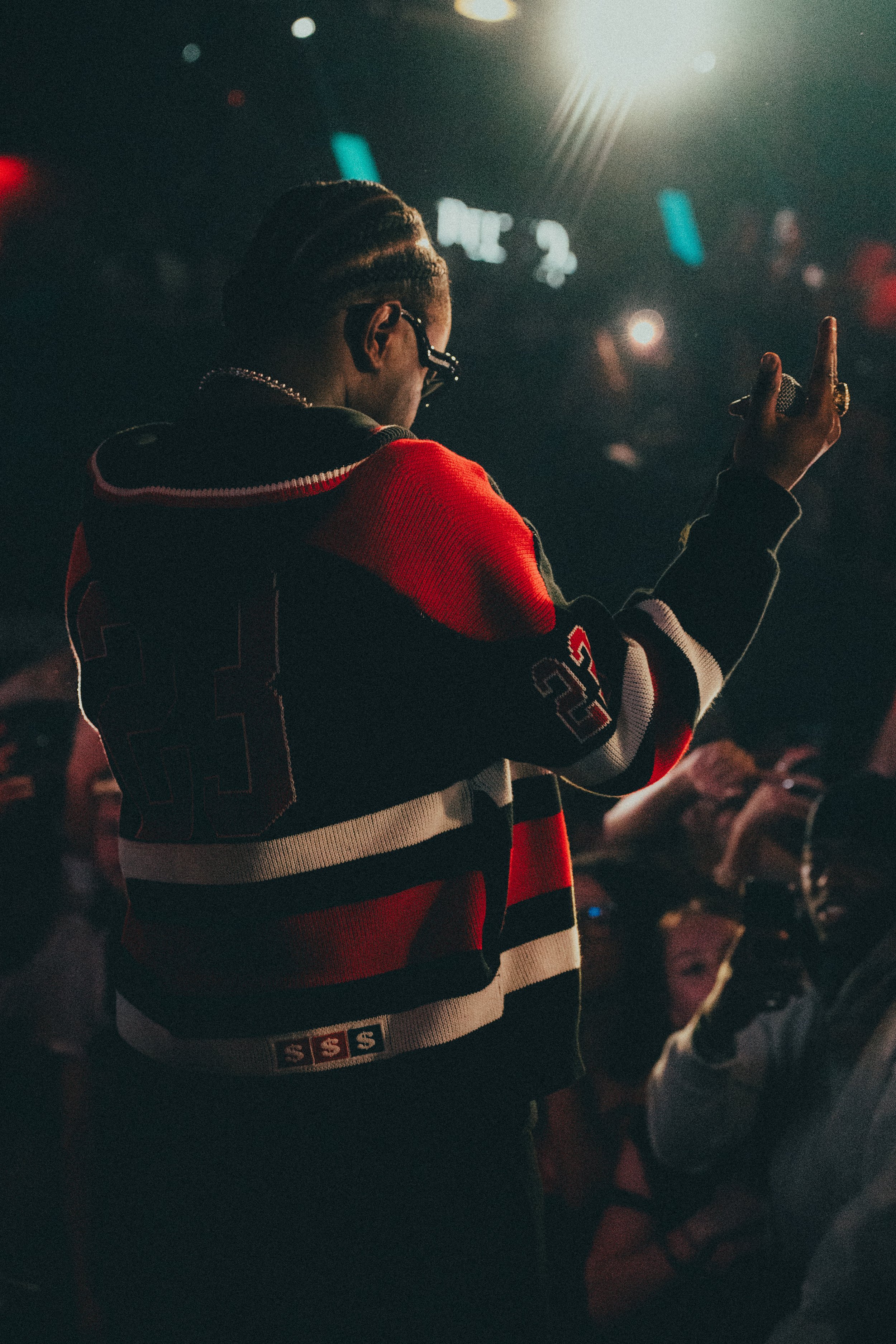 Une personne portant un blouson à rayures rouges, noires et blanches, avec des broderies 93 et un patch 'S' à la taille, fait un geste avec la main dans un lieu sombre avec plusieurs personnes en arrière-plan