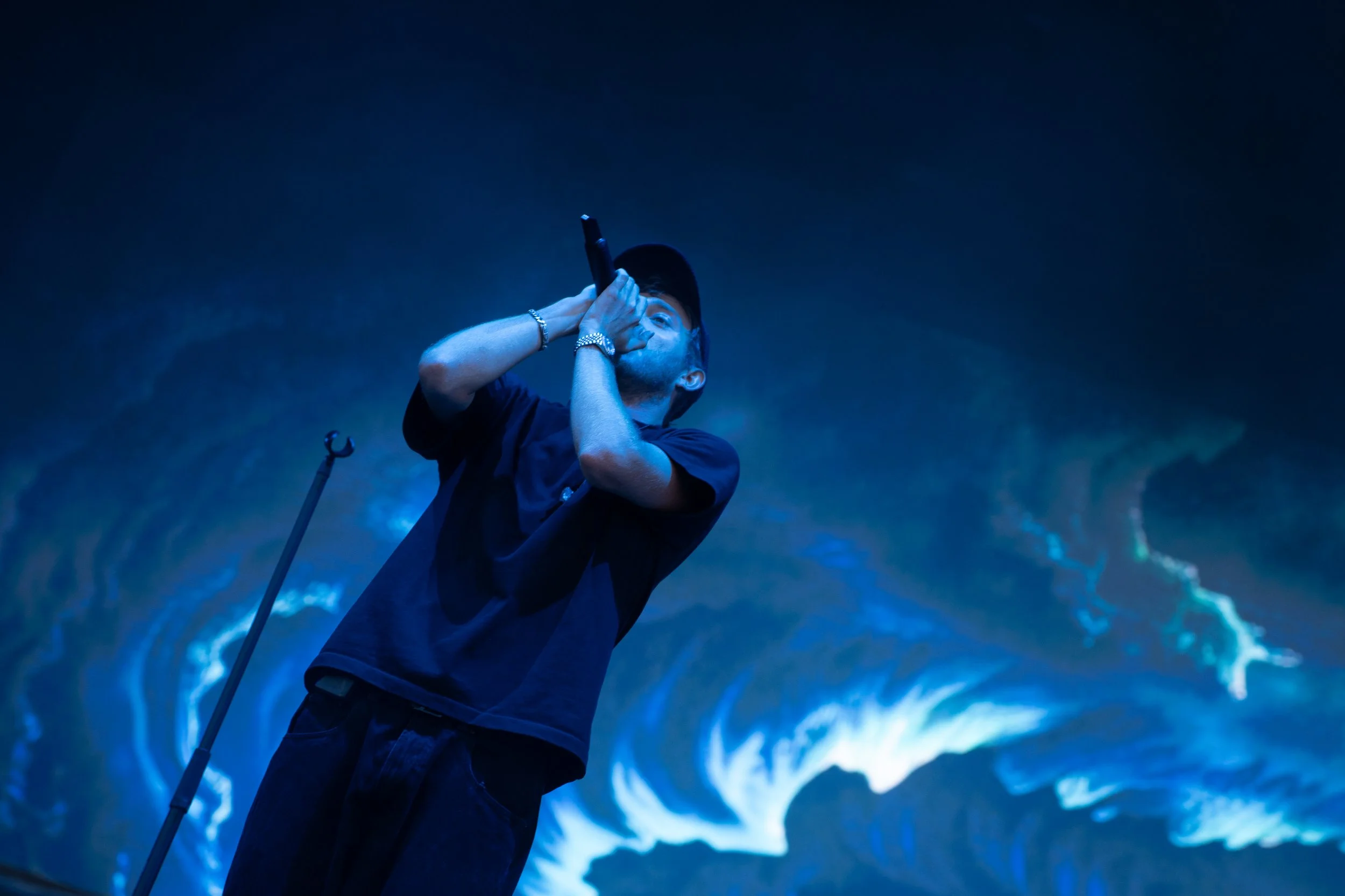 Un jeune homme avec une casquette noire, tenant un microphone, chante ou parle lors d'une performance sur scène avec un fond lumineux bleu et violet évoquant une mer ou un vortex.