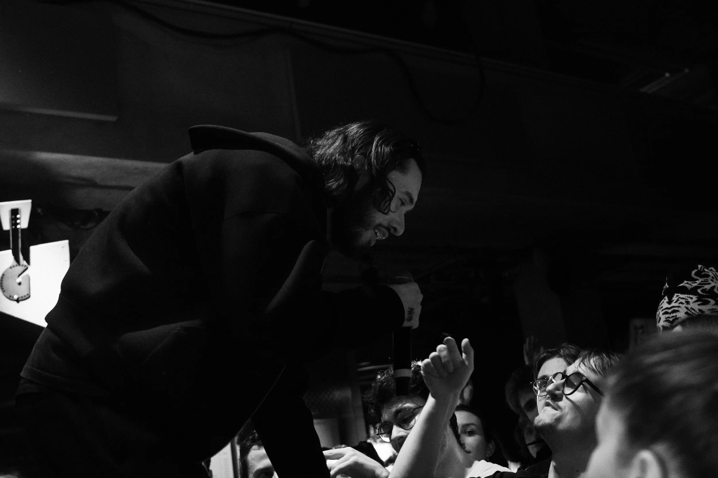 Un homme avec des cheveux longs et une barbe, vêtu d'un hoodie noir, parle ou chante en se tenant penché vers un groupe de personnes plus jeunes, certains portant des lunettes, dans une ambiance sombre.