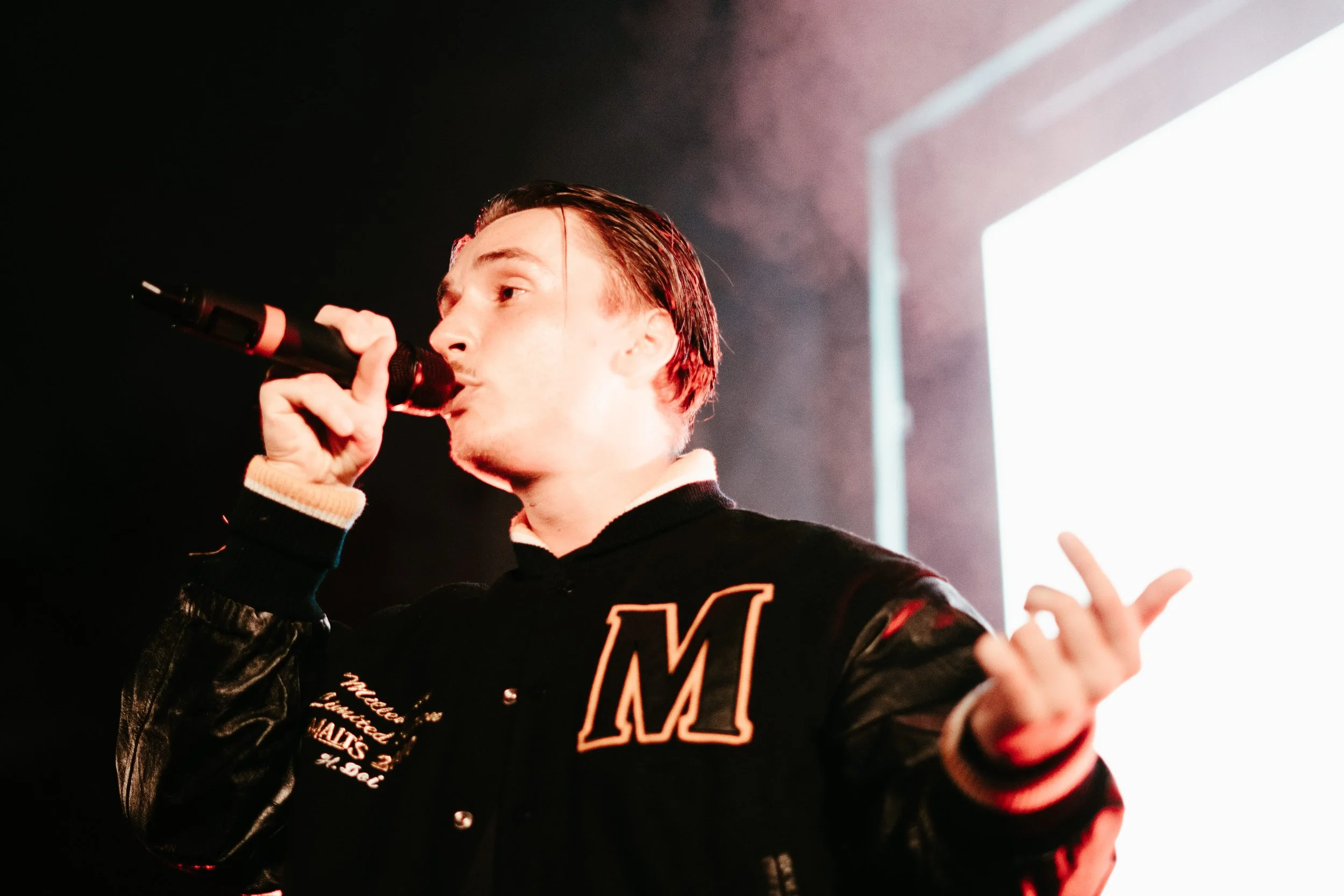 Un jeune homme chante dans un microphone lors d'une performance sur scène, avec un fond lumineux.