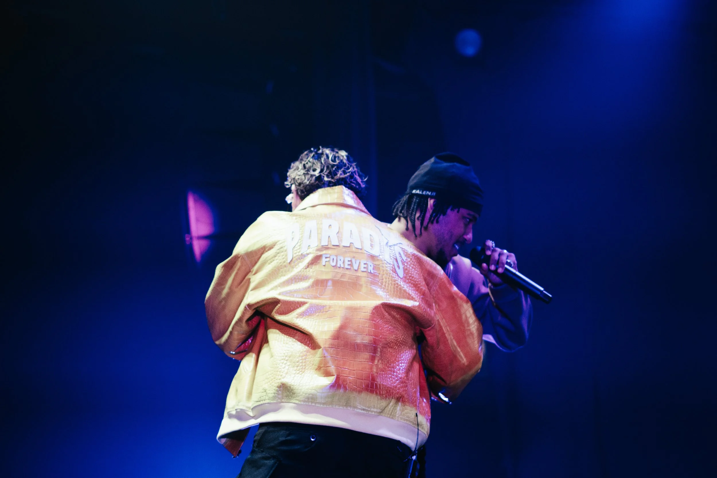 Deux artistes sur scène, l'un de dos portant une veste orange avec 'PARADISE FOREVER' inscrit au dos, et l'autre de face tenant un microphone, sous un éclairage bleu.