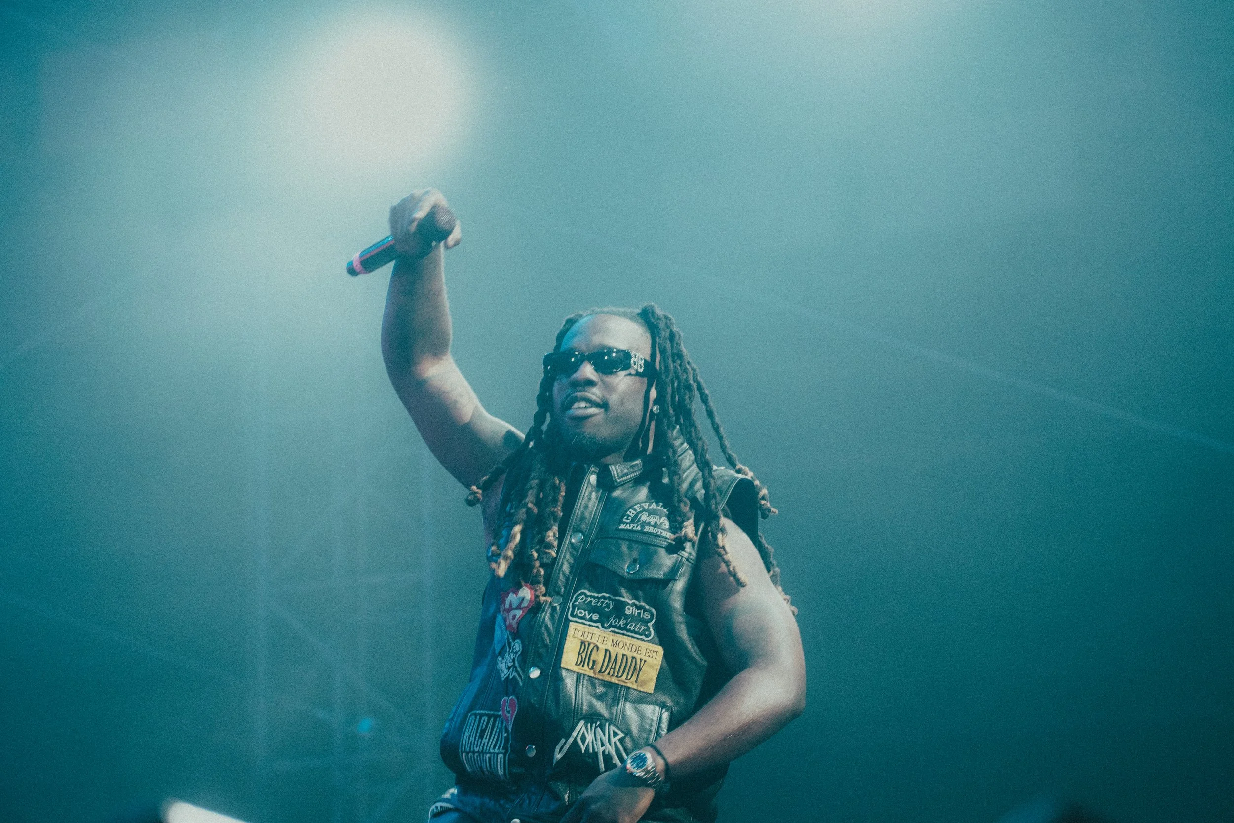 Un homme portant des lunettes de soleil et une veste en cuir décorée de patchs, tenant un microphone en l'air lors d'une performance sur scène.
