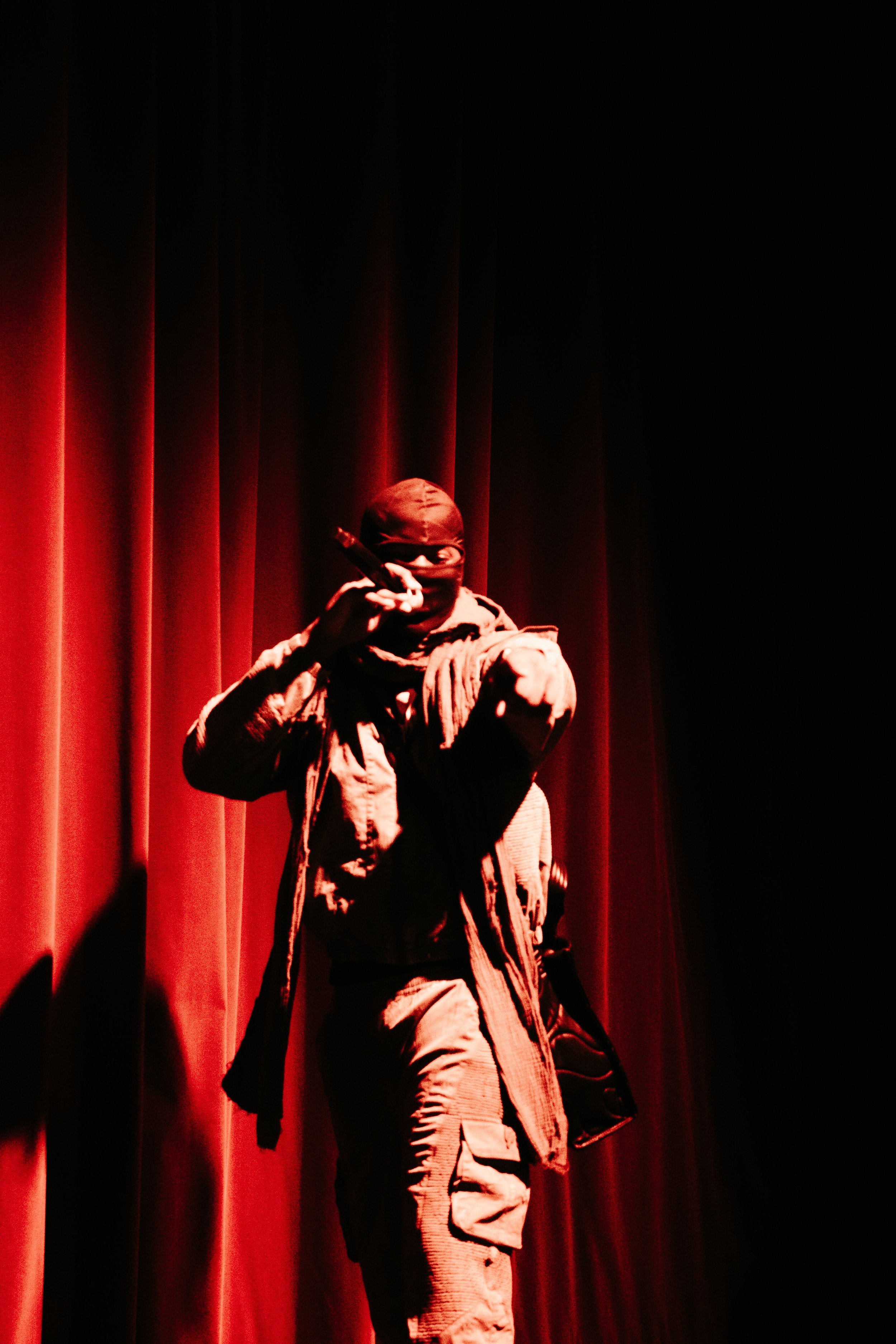 Une personne habillée en noir, portant un casque ou un masque, tenant un micro, debout devant un rideau rouge sur scène, dans une atmosphère sombre