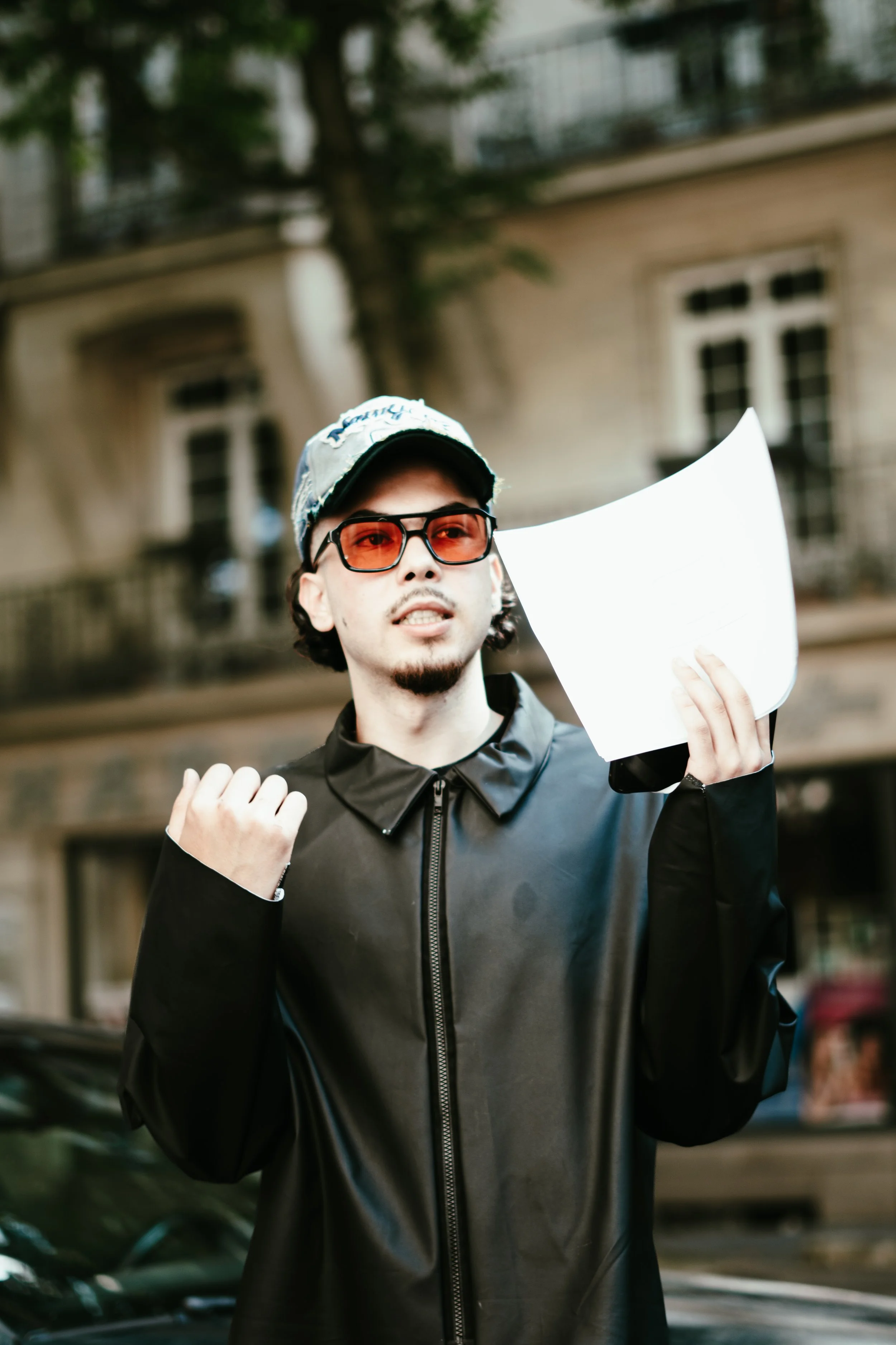 Un homme jeune portant une casquette, des lunettes de soleil oranges, une veste noire en cuir, tenant un papier blanc dans sa main droite, levant légèrement le poing gauche, dans une rue urbaine.
