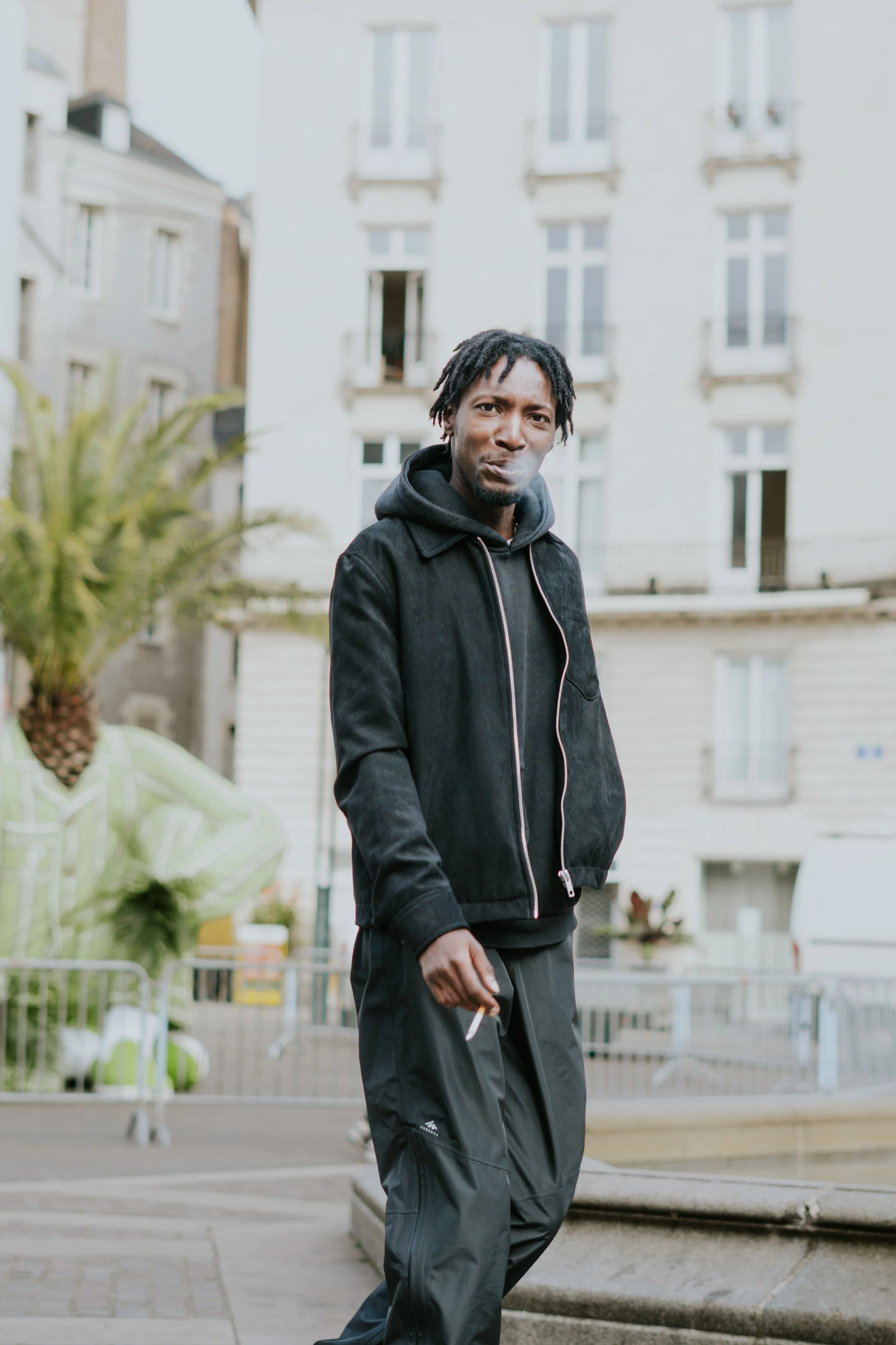 Un homme portant une veste noire et un sweat à capuche noir, debout dans une rue urbaine, fumant une cigarette.