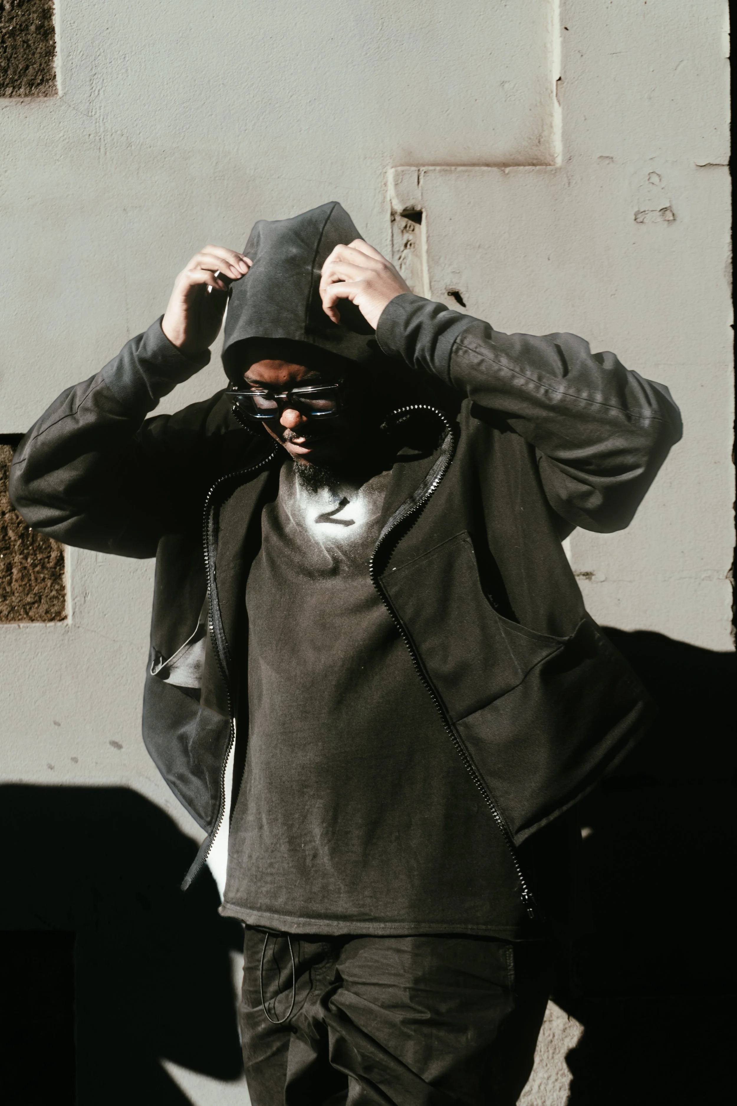 Homme en vêtements noirs, portant une cagoule et des lunettes de soleil, se tenant contre un mur en écoutant de la musique.
