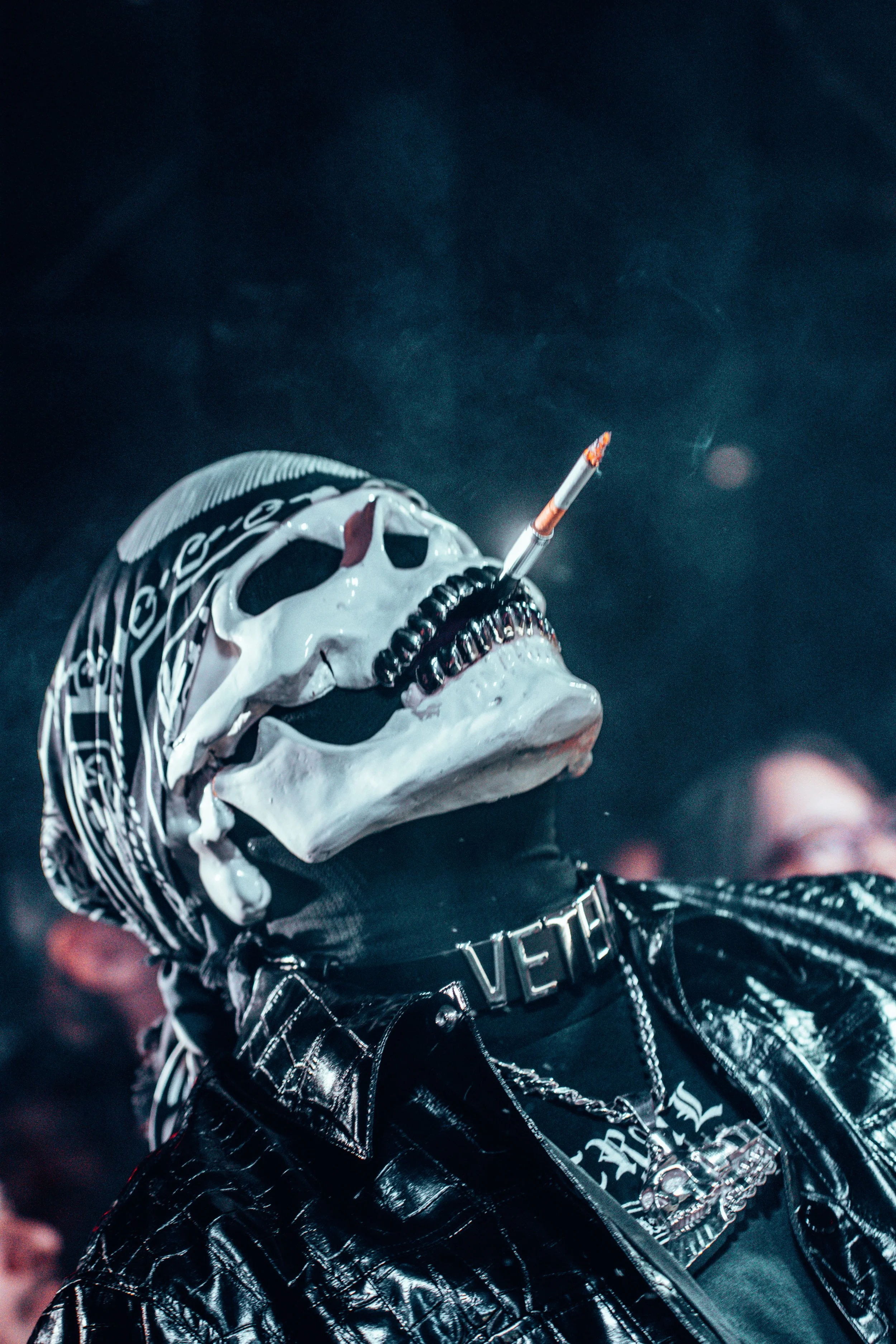 Personne portant un masque de crâne avec un cigarette à la bouche, vêtue de vêtements en cuir noirs, dans une ambiance sombre et mystérieuse.
