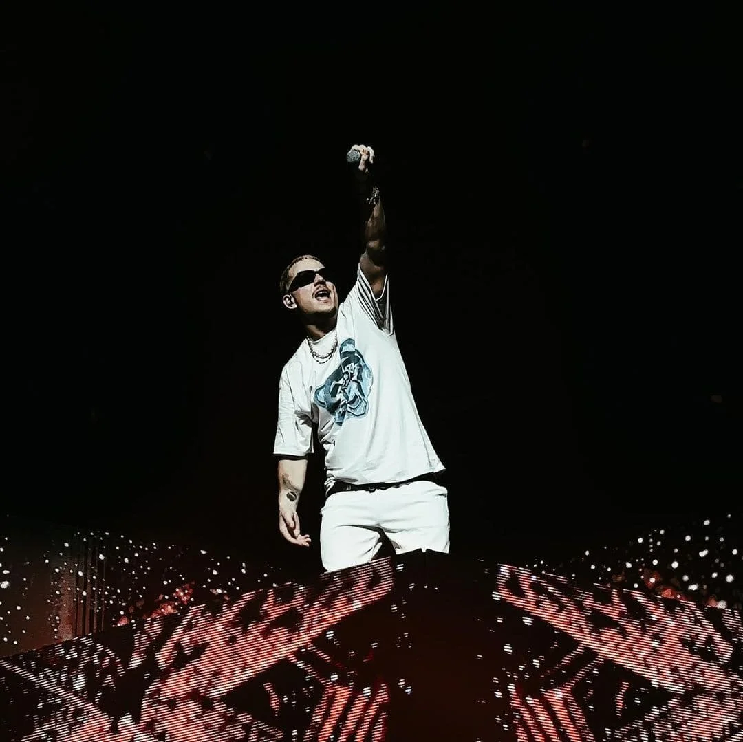 PLK portant des lunettes noires, un t-shirt blanc avec une illustration bleue, et des bijoux, fait un geste en l'air lors d'un concert ou d'un spectacle, avec un fond noir.