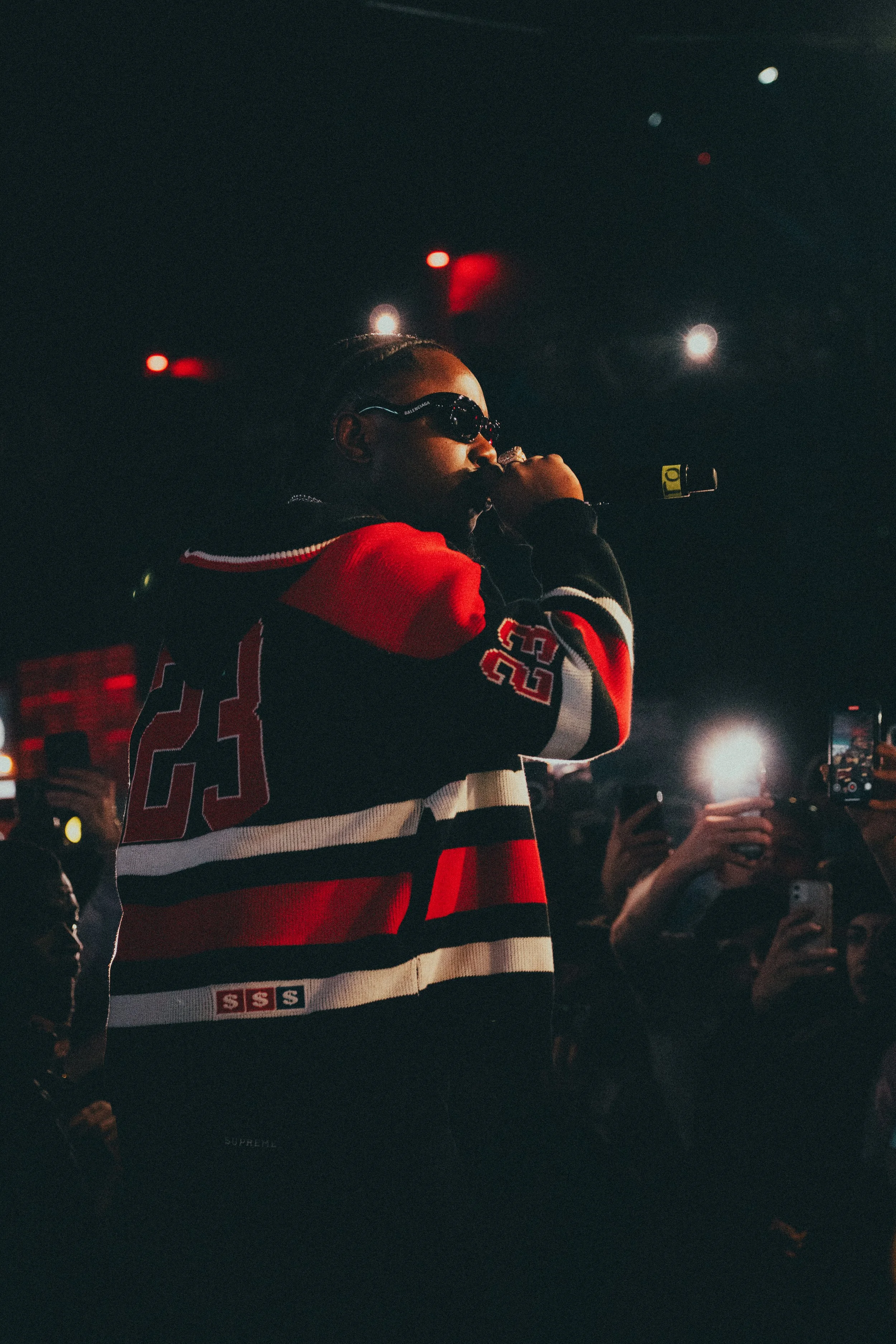 Un artiste en train de rapper avec des lunettes de soleil, portant un sweat à rayures rouges, noirs et blancs, sur scène lors d'un concert, entouré d'un public qui le filme avec des téléphones.