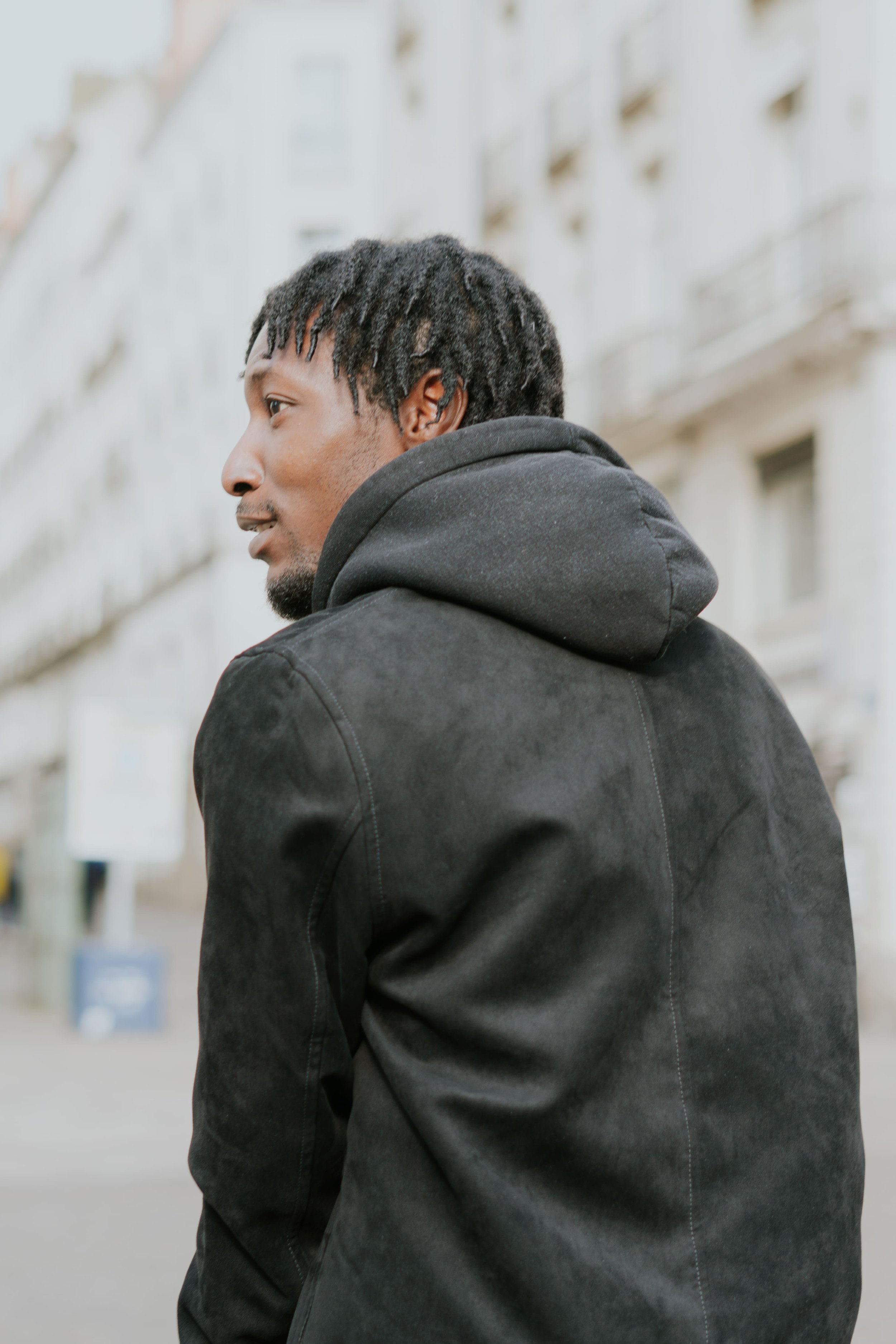 Homme avec des dreadlocks portant une veste noire et un sweat à capuche gris, vue de profil dans une rue urbaine.