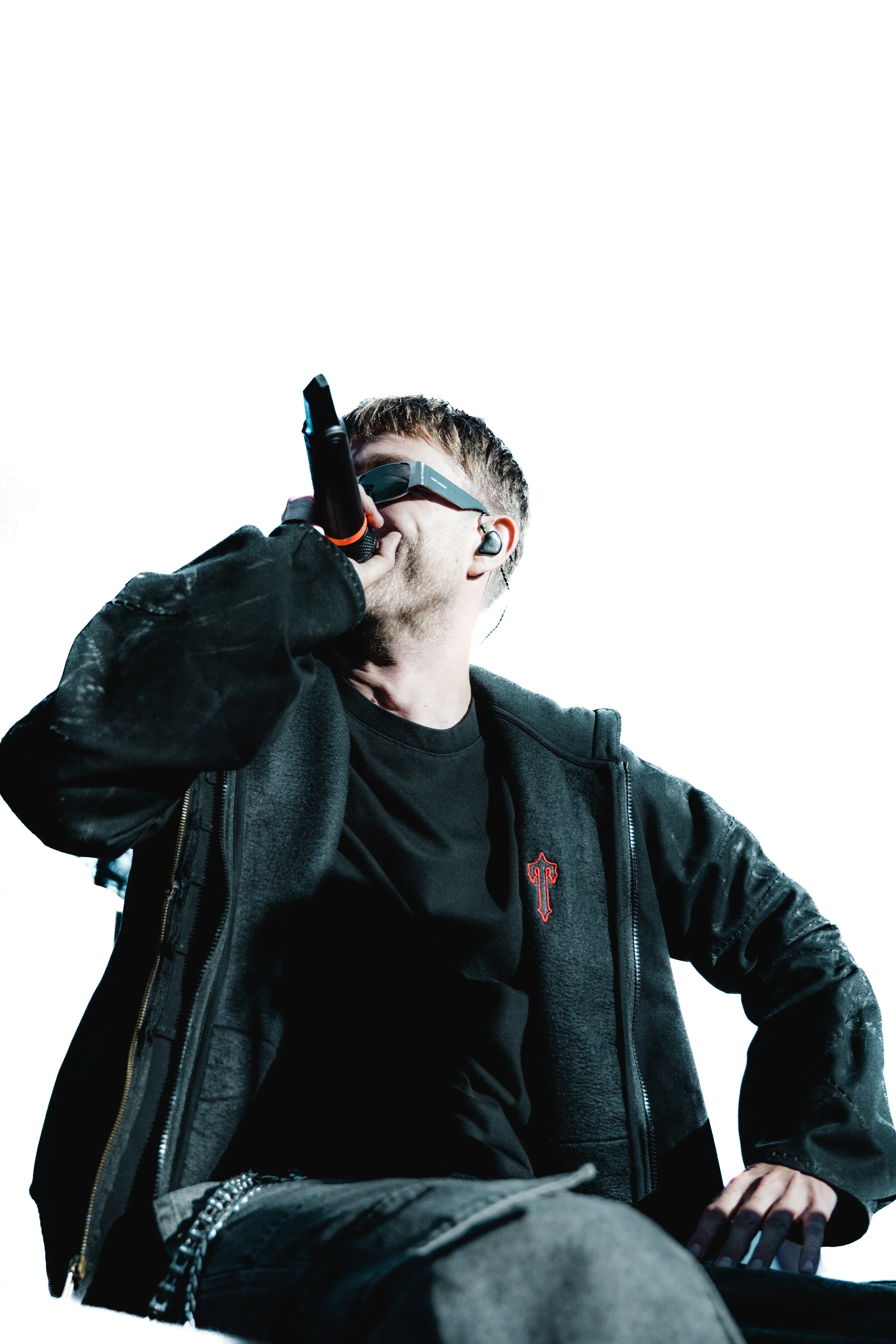 Un chanteur ou rappeur portant des lunettes de réalité virtuelle, tenant un microphone, vêtu d'un hoodie noir avec un logo rouge, sur scène.