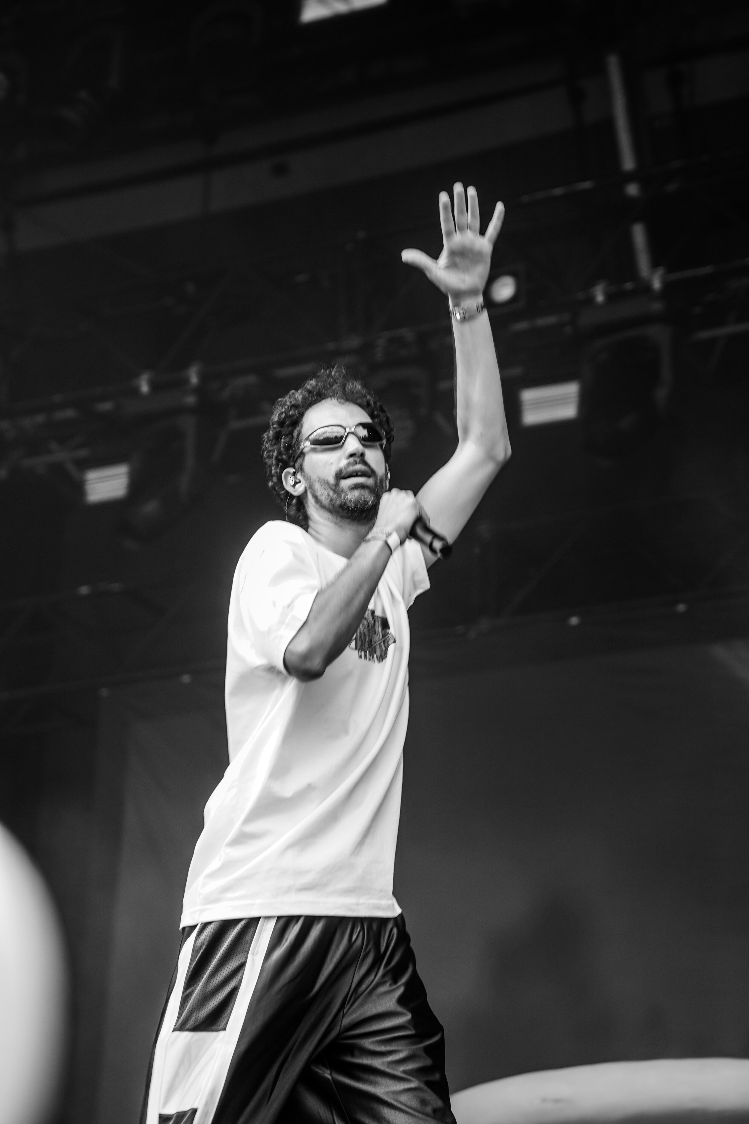 Homme avec barbe et cheveux bouclés portant des lunettes de soleil, un t-shirt blanc et un pantalon noir, parlant dans un microphone sur scène, au concert.